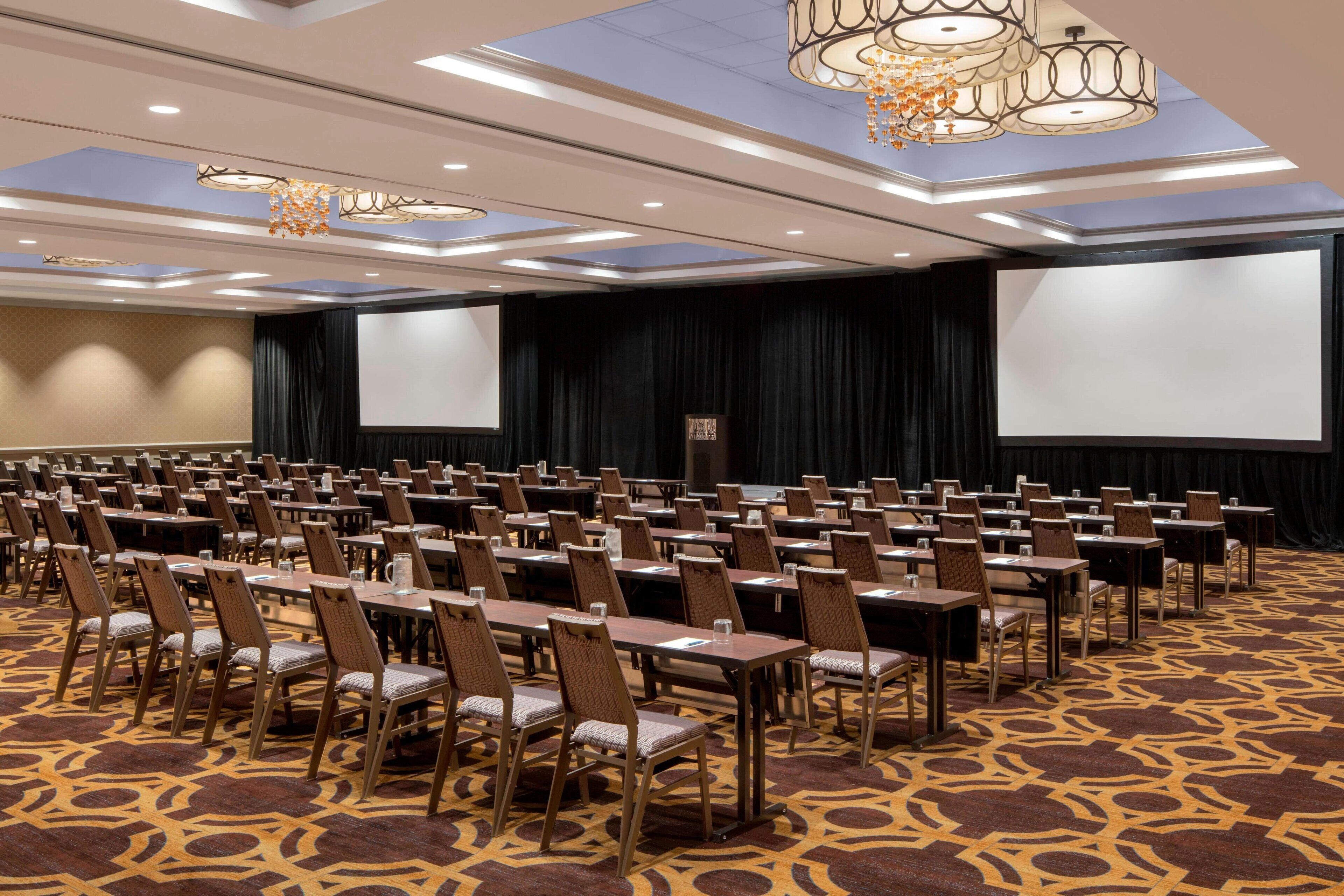 A conference room with rows of tables and chairs, large screens, and chandeliers.
