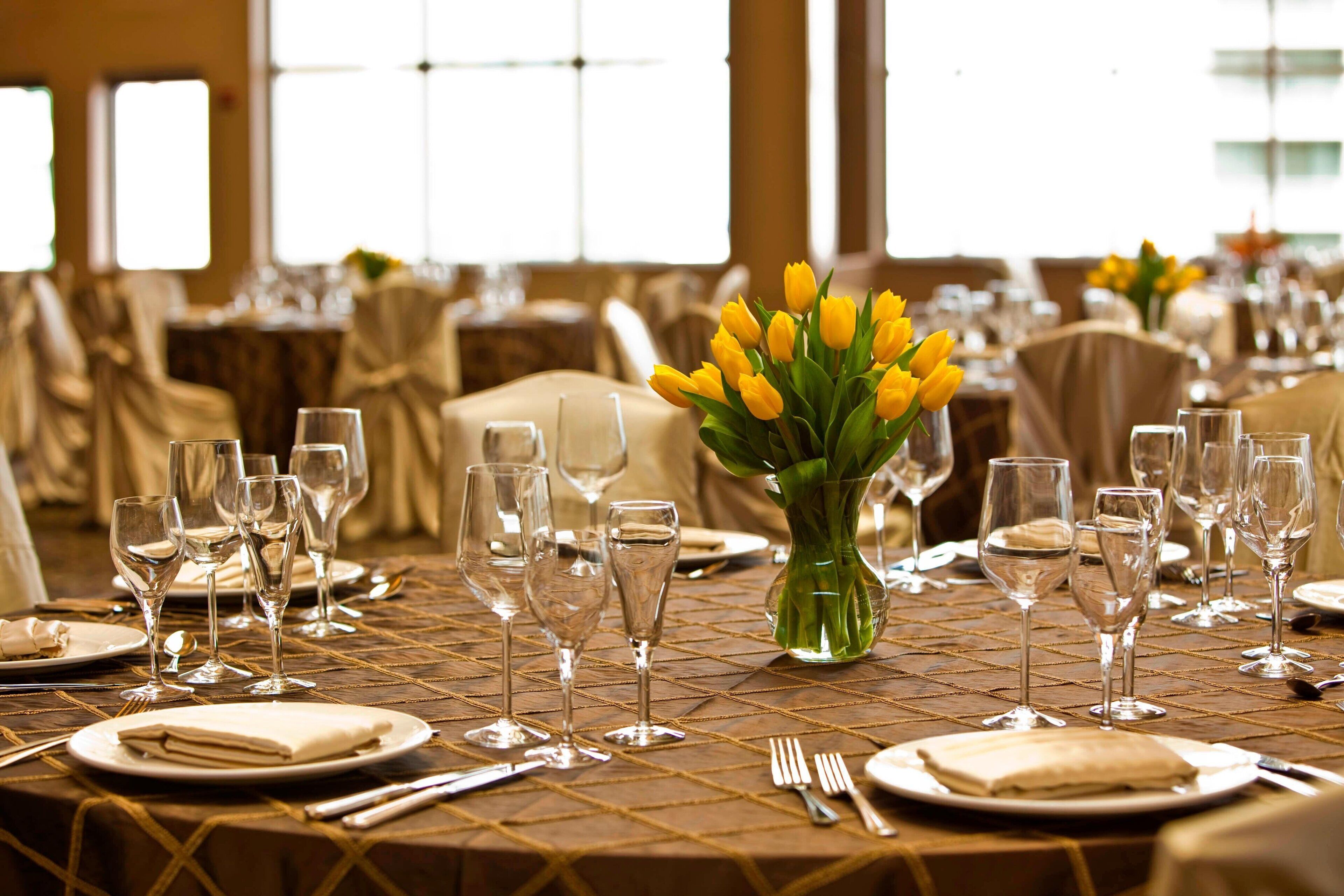 A banquet table set with wine glasses, plates, and a vase of yellow tulips.