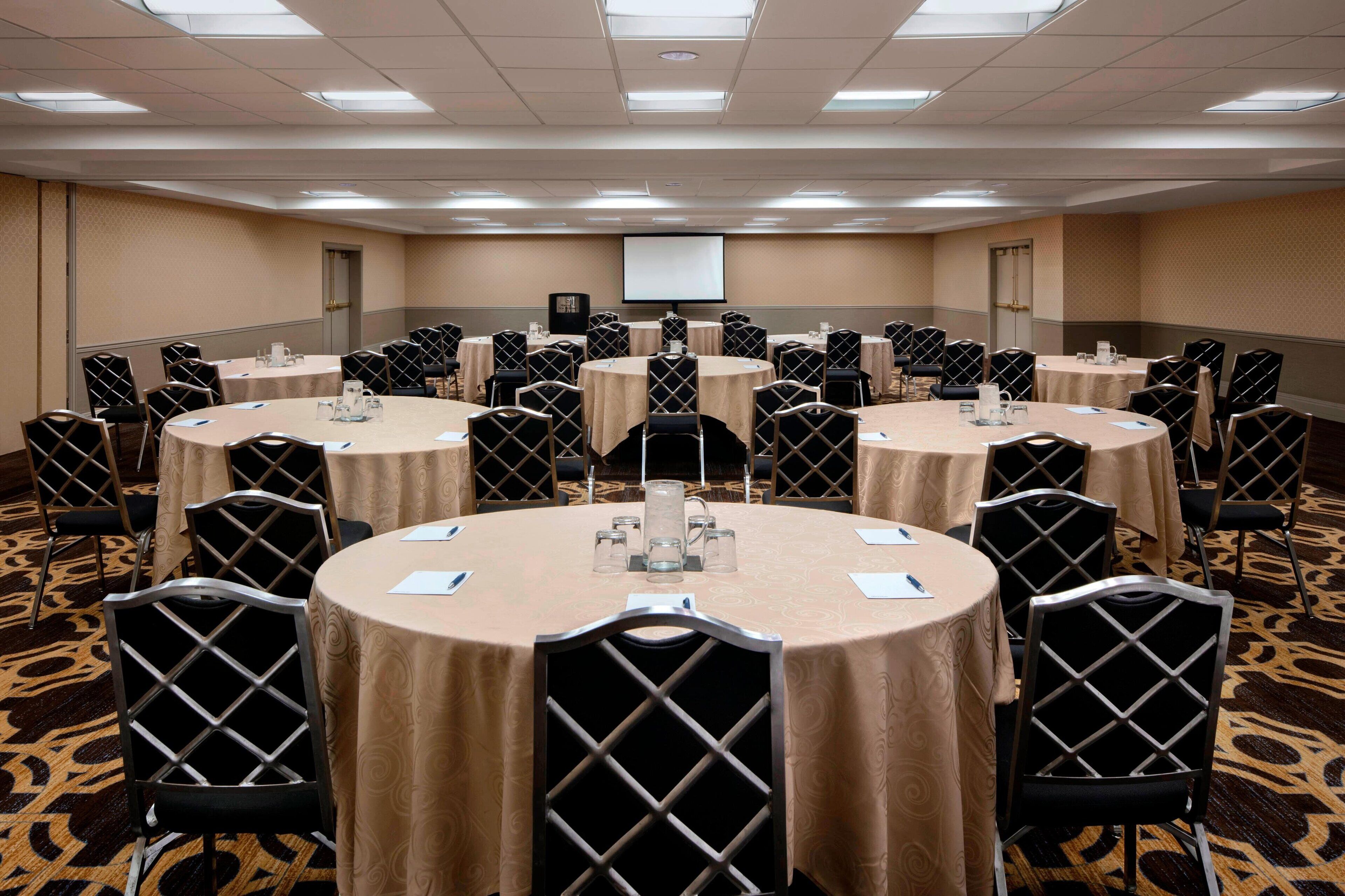 A conference room with multiple round tables, each surrounded by chairs with a diamond-patterned backrest. Tables are covered with beige tablecloths and have water pitchers and notepads on them.