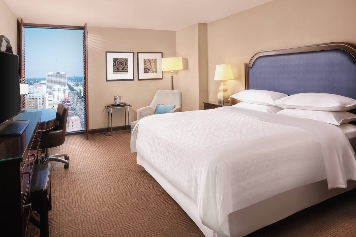 A hotel room with a large bed, a desk with a chair, a small table with a lamp, and a view of the city through a large window.