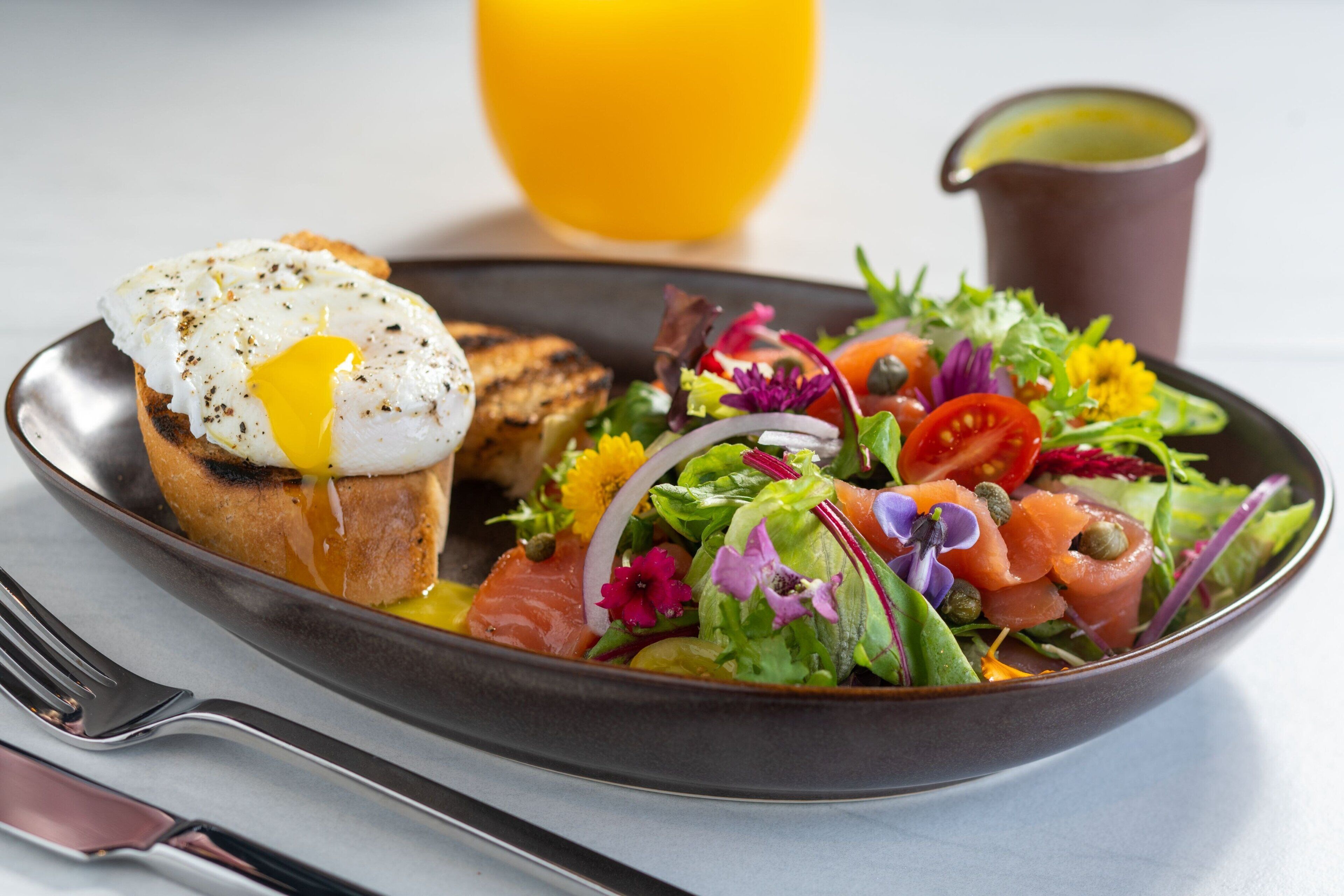 A breakfast plate with a poached egg on toast, a salad with various vegetables and salmon, a glass of orange juice, and a small pitcher of sauce.