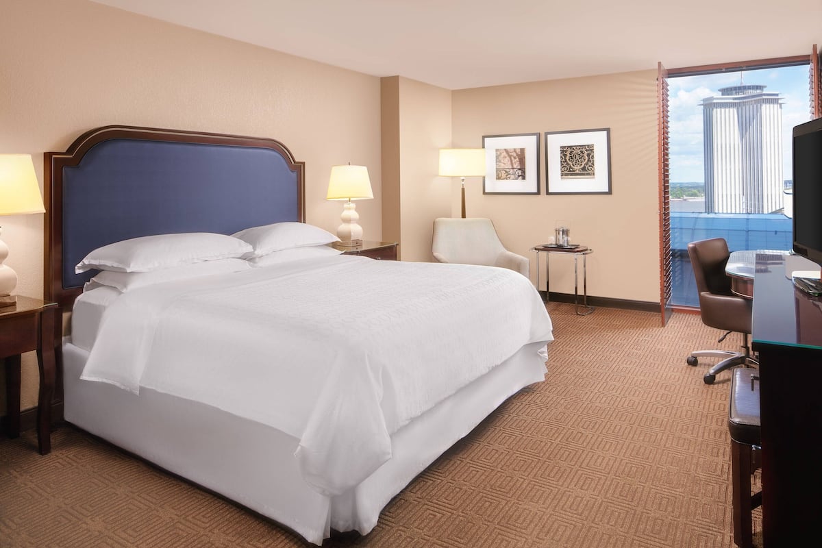 A hotel room with a large bed, two bedside lamps, a desk with a chair, a television, and framed artwork on the wall.