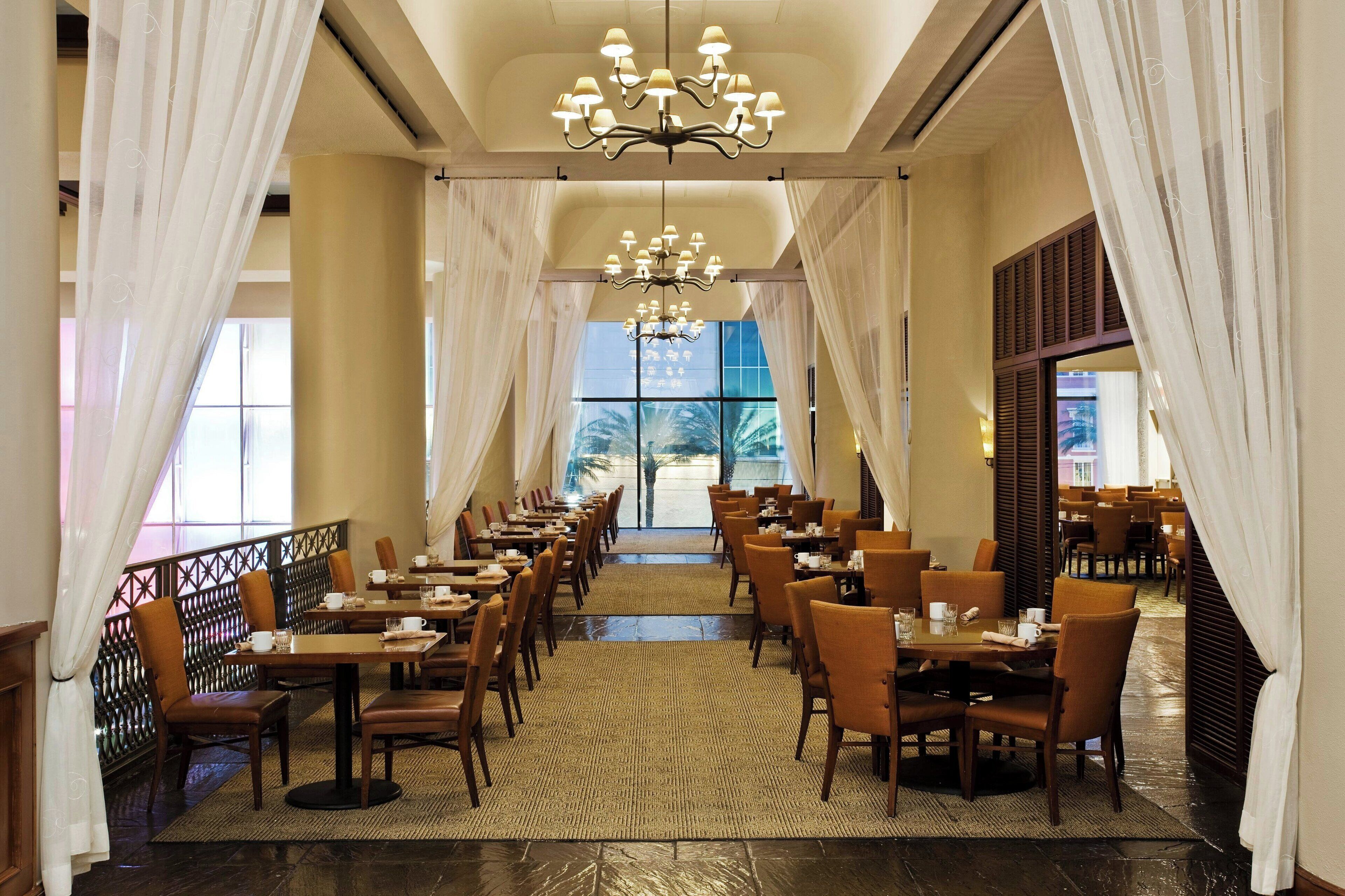 A spacious dining area with multiple tables and chairs, a chandelier, and large windows allowing natural light.