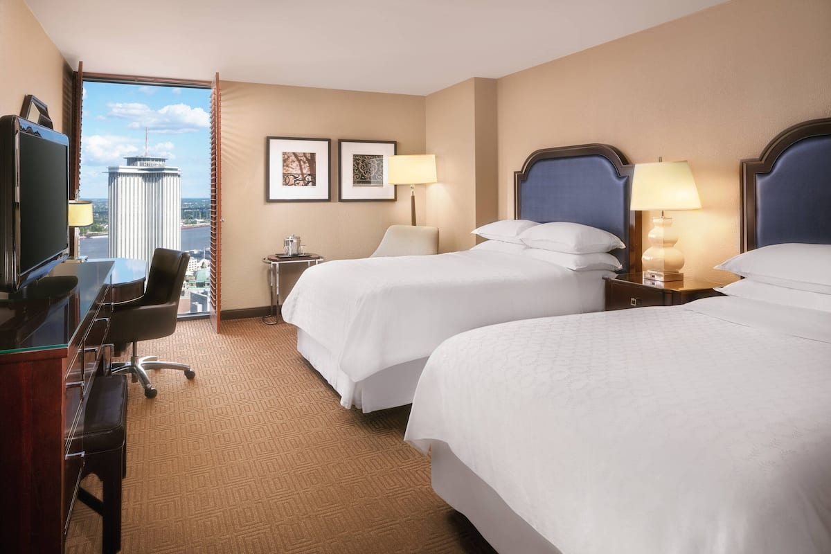 A hotel room with two beds, a desk with a chair, a TV, and a view of a tall building.