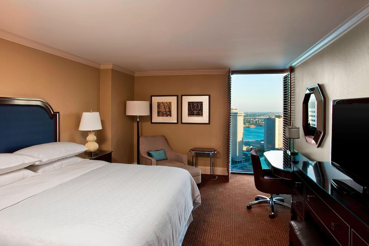 A hotel room with a large bed, a desk, a chair, a TV, and a view of the city.