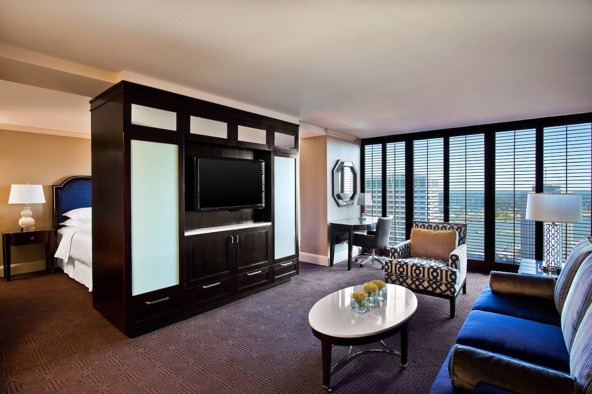 A hotel room with a large flat-screen TV, a sofa, a coffee table with flowers, a desk, and a chair.