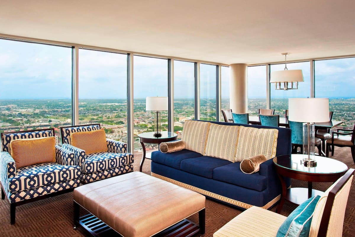 A high-rise hotel room with a large city view, a blue sofa, patterned armchairs, and a round table with a lamp.