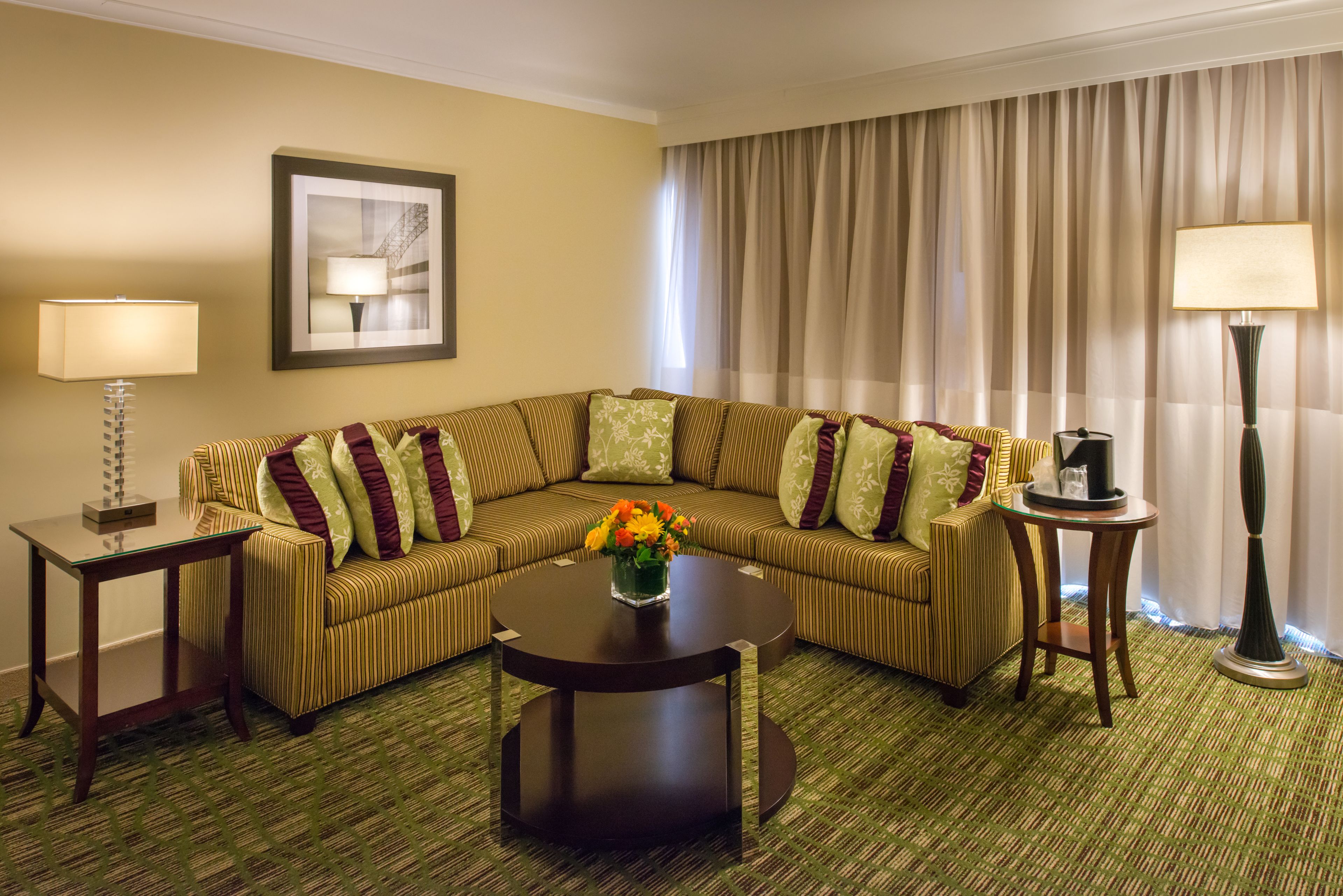 A hotel room with a striped sofa, a round coffee table with a vase of flowers, two side tables with lamps, and a large window with curtains.