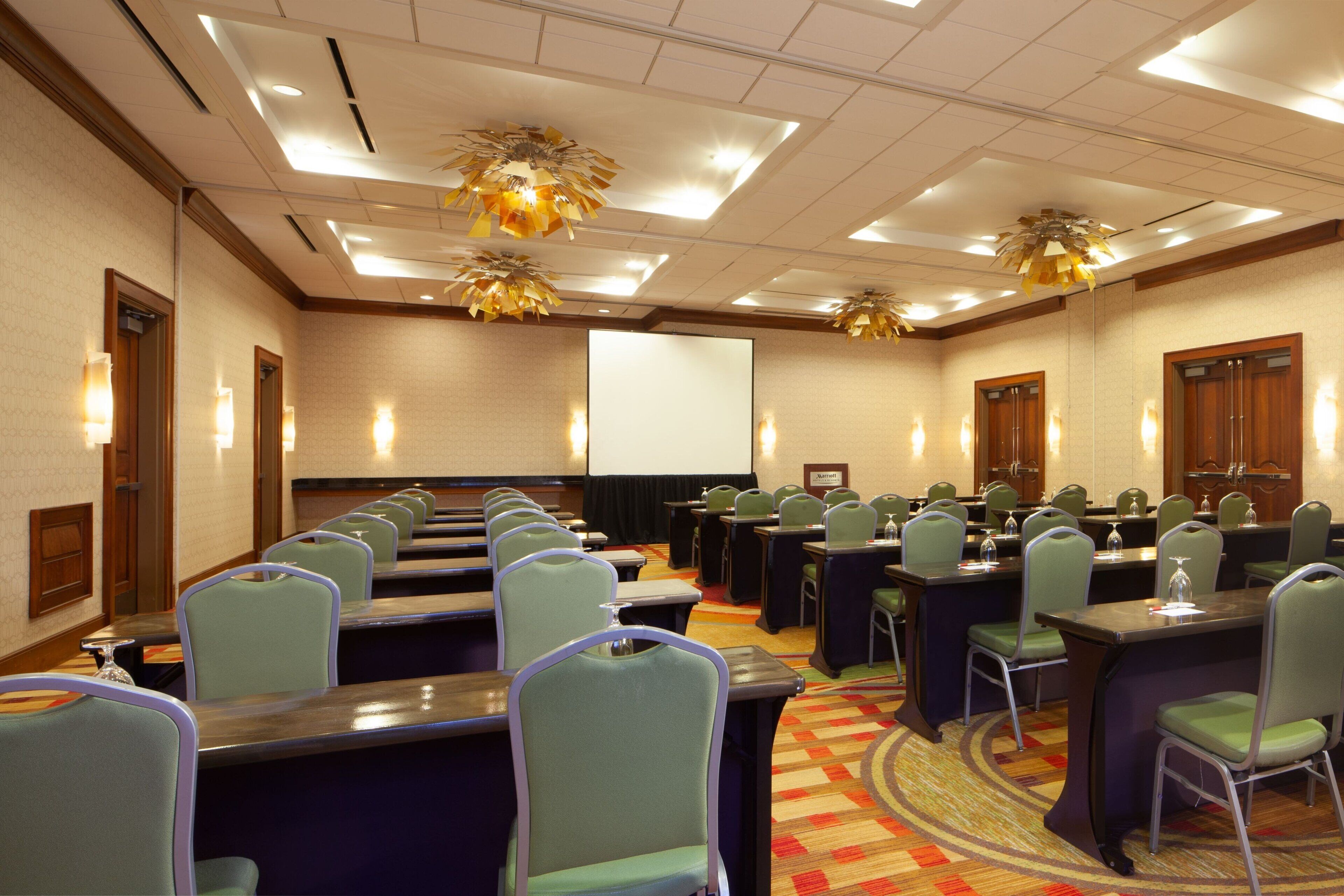 A conference room with rows of tables and chairs, a projector screen, and ceiling lights.