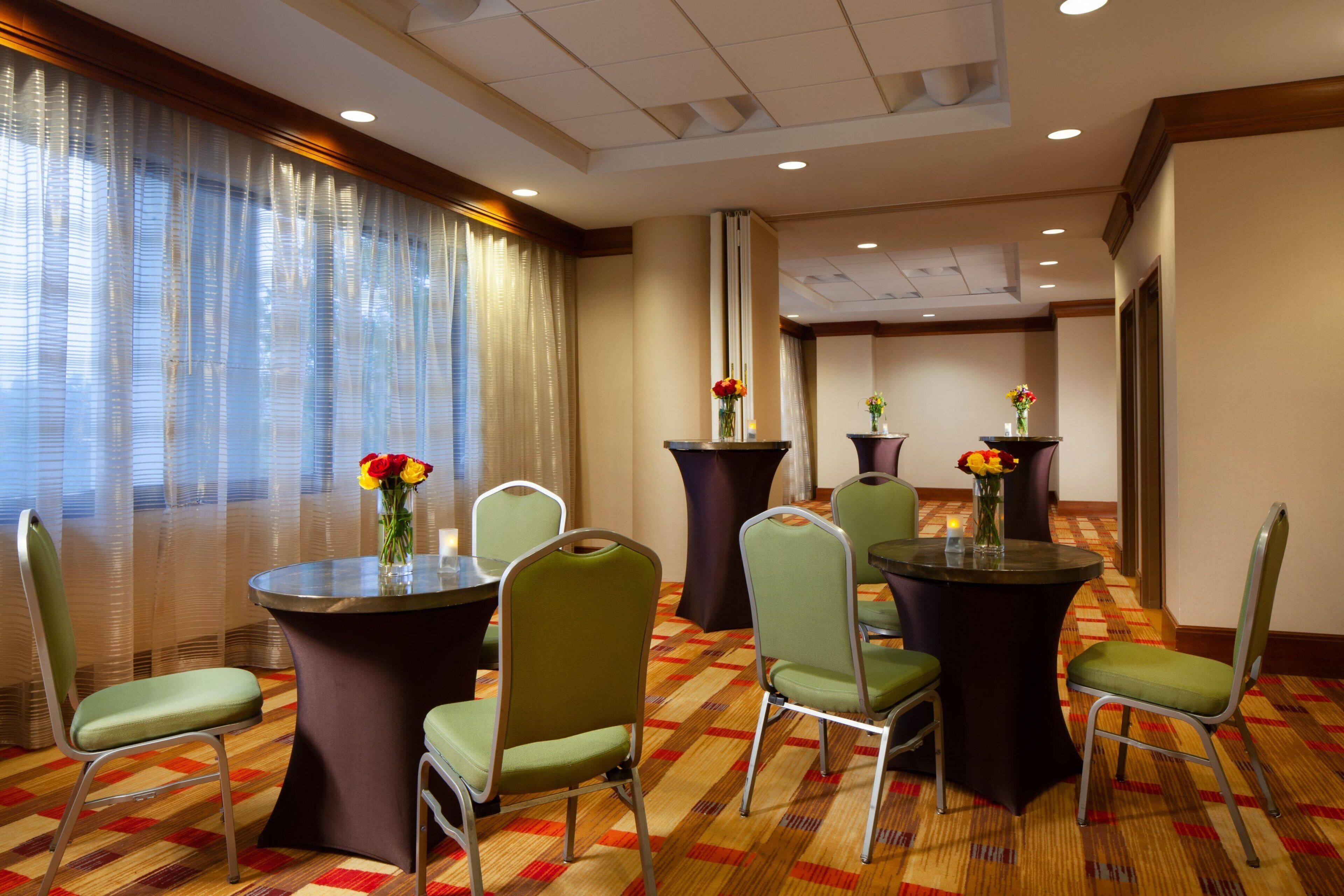 Une chambre d’hôtel dotée d’une grande fenêtre, d’un sol recouvert de moquette et d’un coin repas avec des tables et des chaises rondes.
