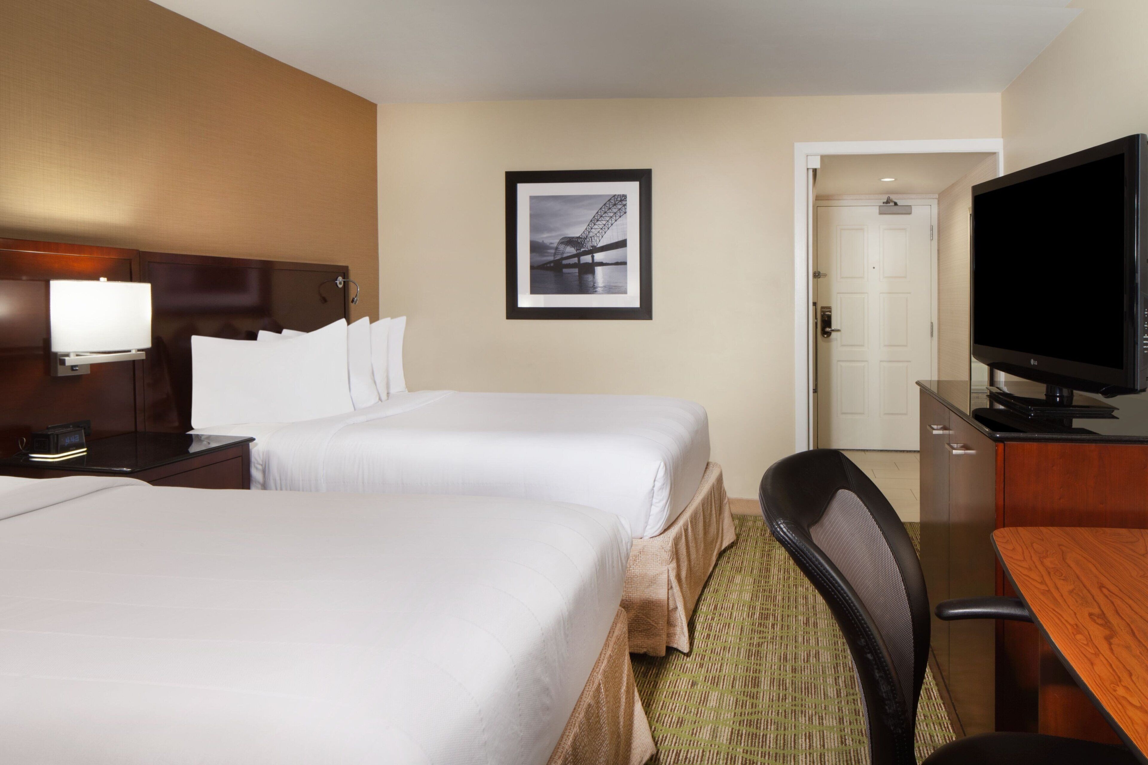 A hotel room with two beds, a desk with a television, a framed picture on the wall, and a door leading to another room.