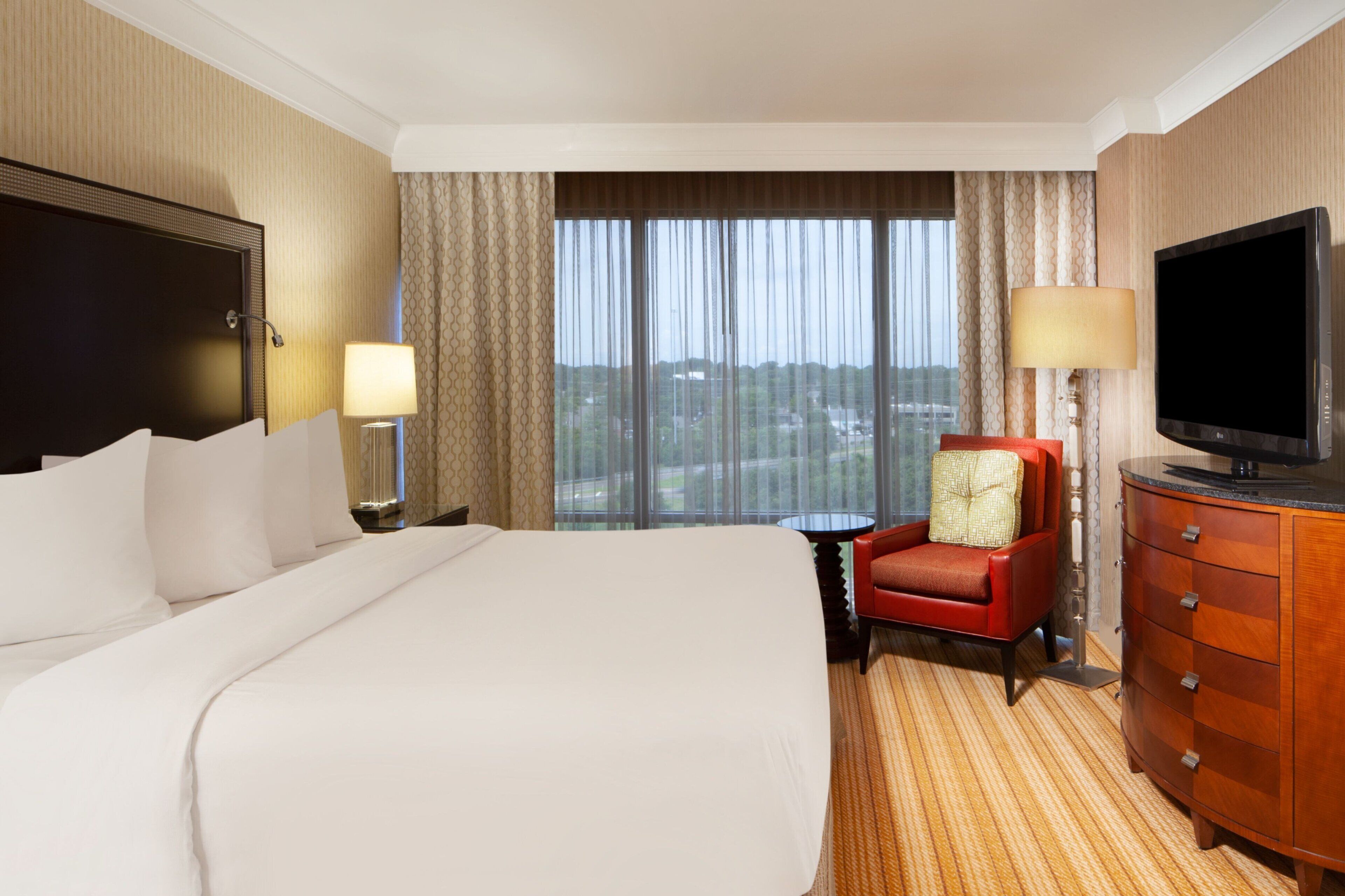 Ein Hotelzimmer mit einem großen Bett, einem Fernseher, einem roten Sessel und Blick auf die Stadt durch das Fenster.
