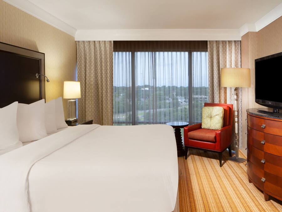 Habitación de hotel con una cama grande, televisión, un sillón rojo y vista a la ciudad por la ventana.