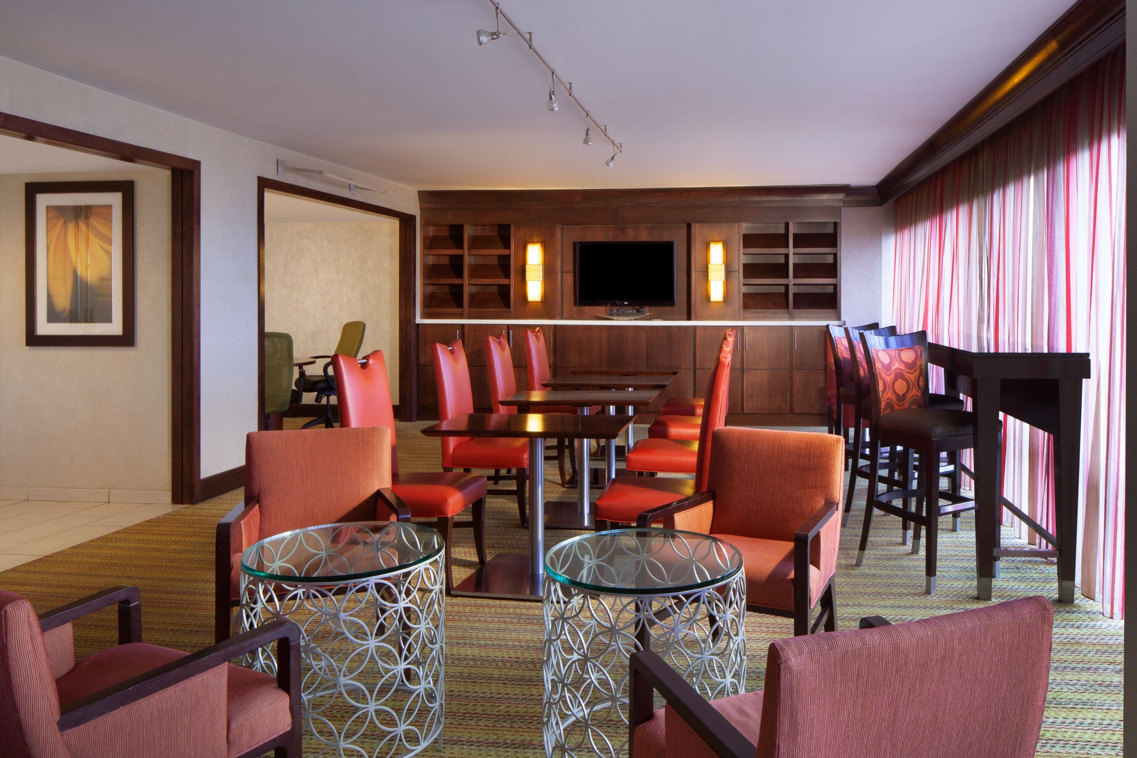 Habitación de hotel con zona de comedor, televisión y un gran ventanal con cortinas.