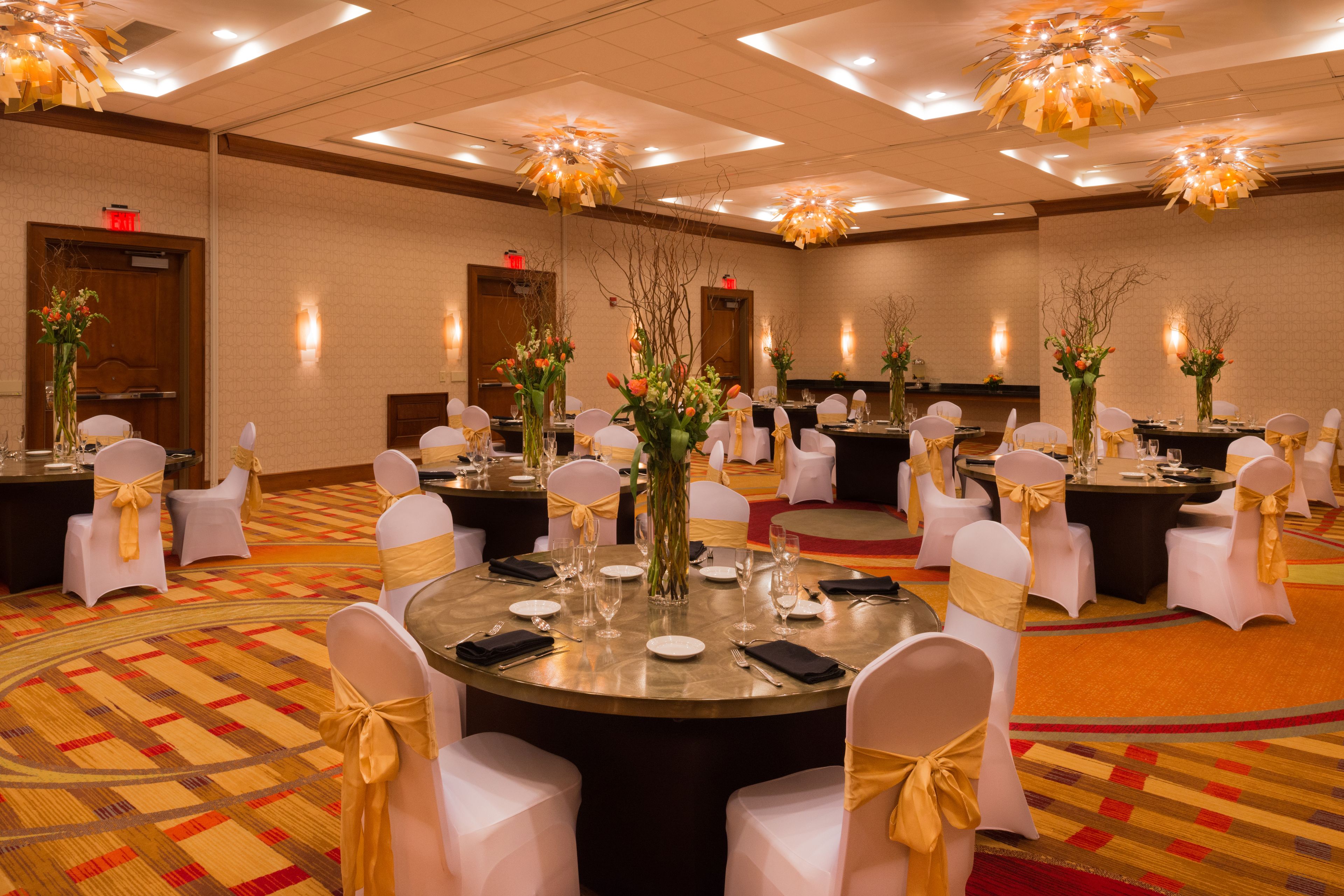 A banquet hall with round tables, white chairs with gold sashes, and floral centerpieces.