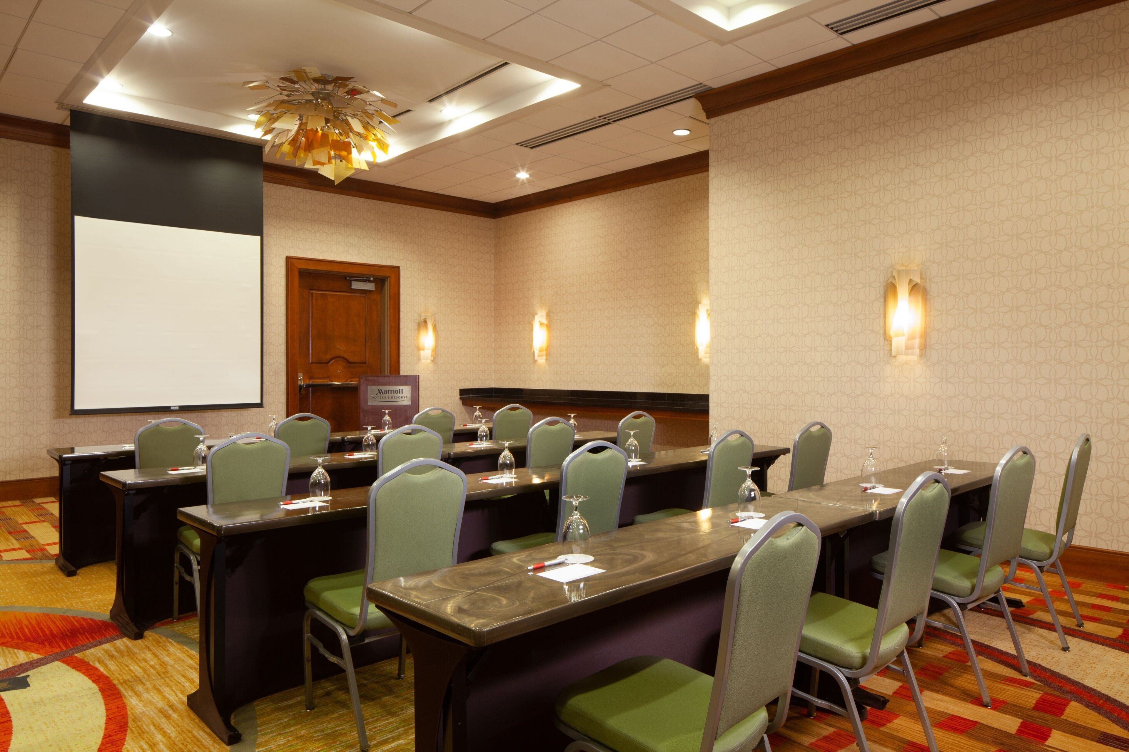 A conference room with a long table, multiple chairs, a projector screen, and a carpeted floor.