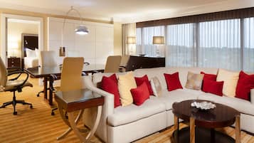 A hotel room with a large L-shaped sofa, a dining table, a desk, and a large window with curtains.