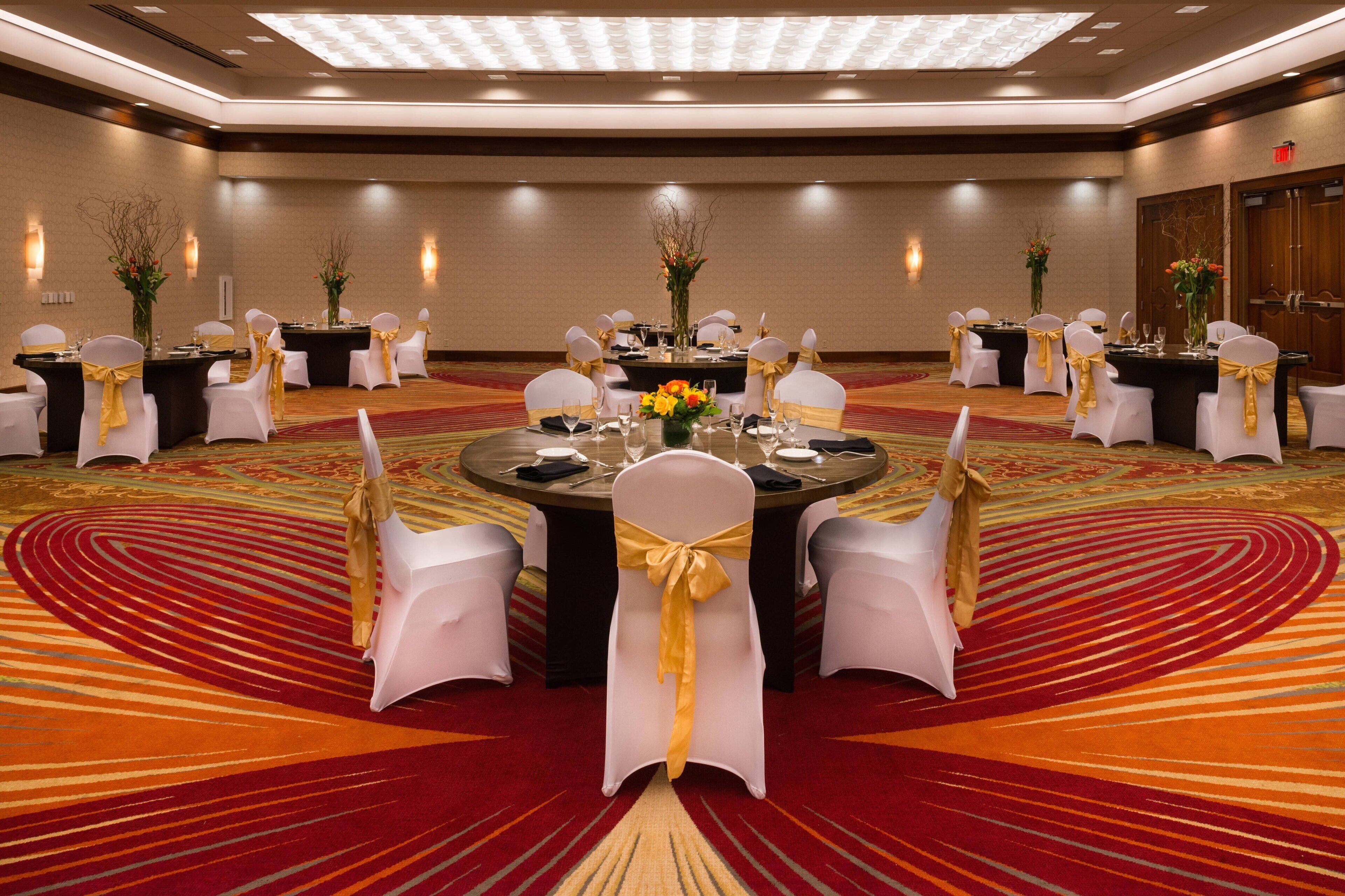 Un salón de conferencias con mesas redondas, sillas blancas con cintas doradas y centros de mesa florales.