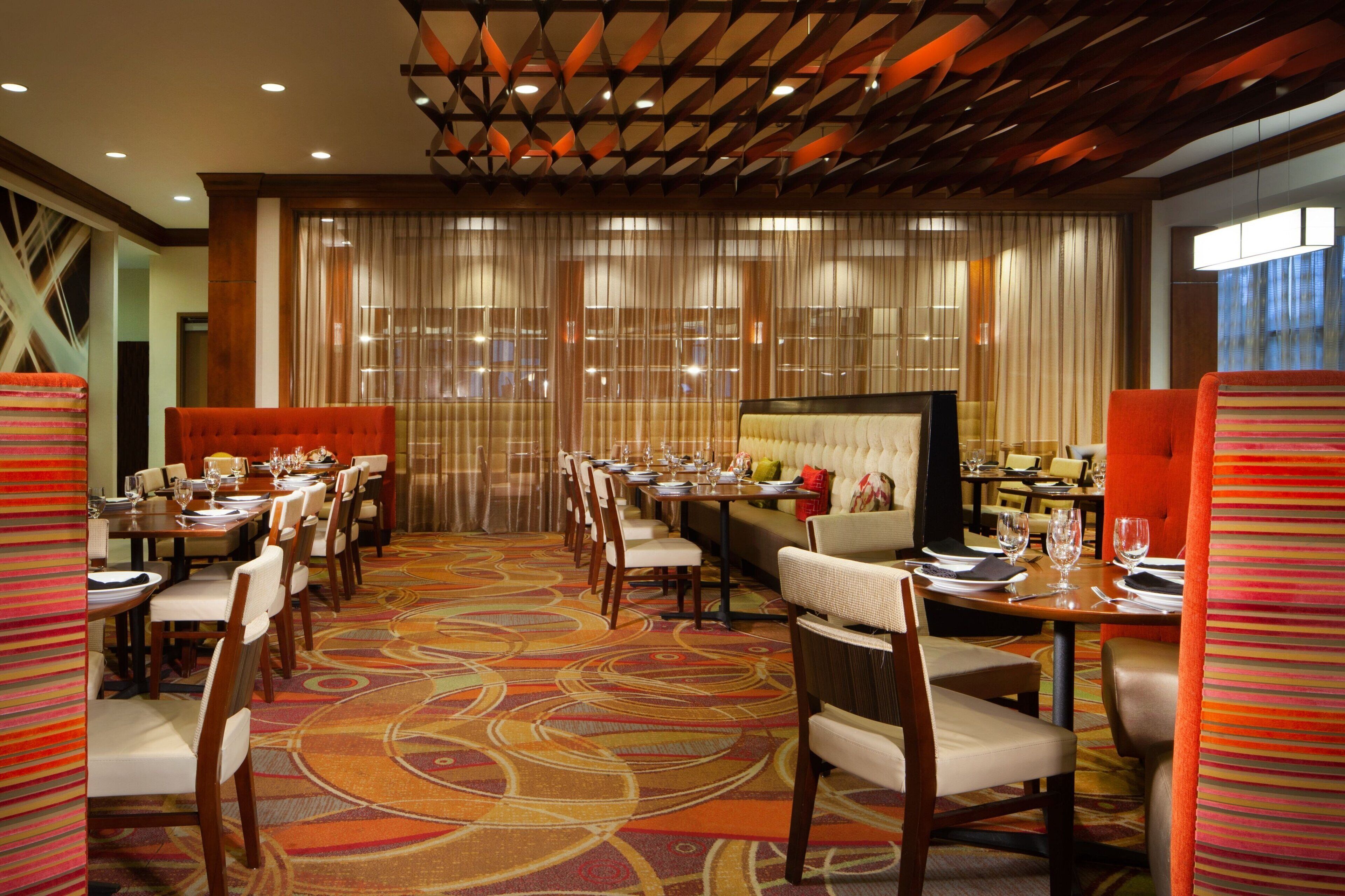 A hotel dining area with multiple tables and chairs, a patterned carpet, and a modern ceiling design.