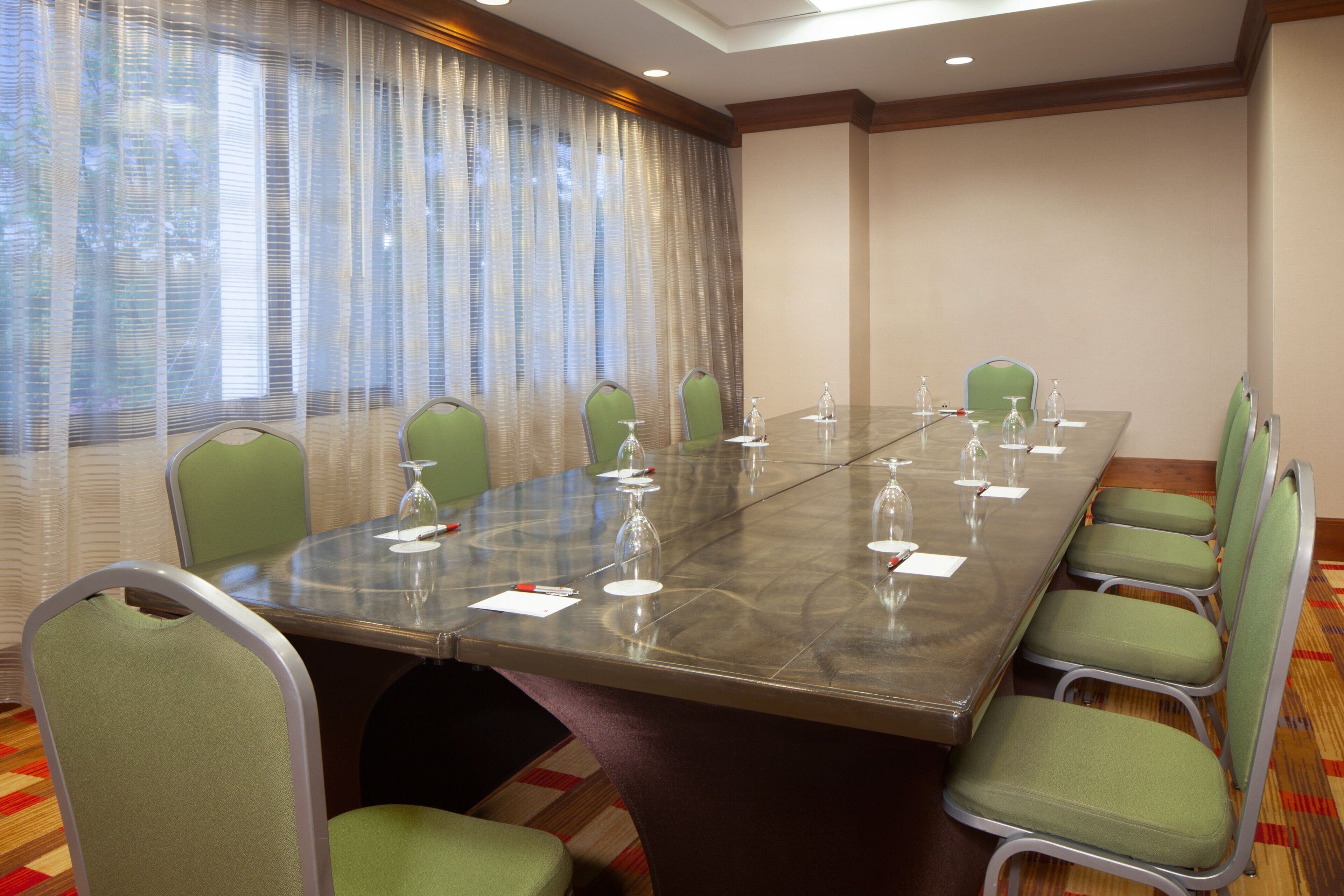 A conference room with a glass table, green chairs, and a window with sheer curtains.