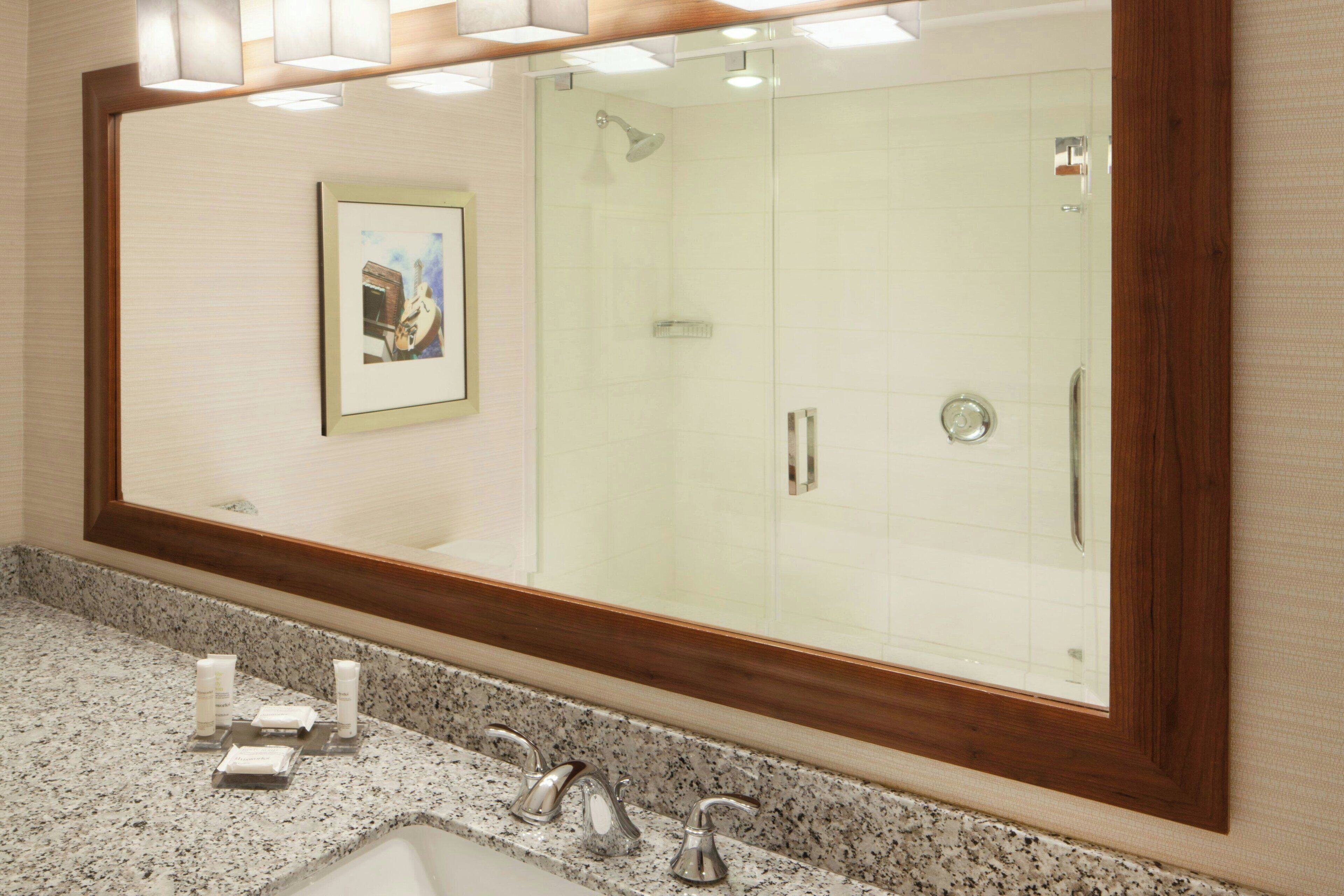 A bathroom with a large mirror, a framed picture on the wall, a granite countertop, and a shower area.