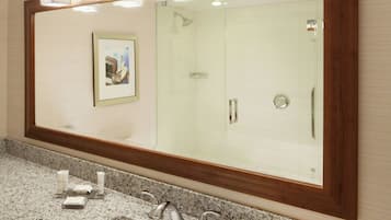 A bathroom with a large mirror, a framed picture on the wall, a granite countertop, and a shower area.