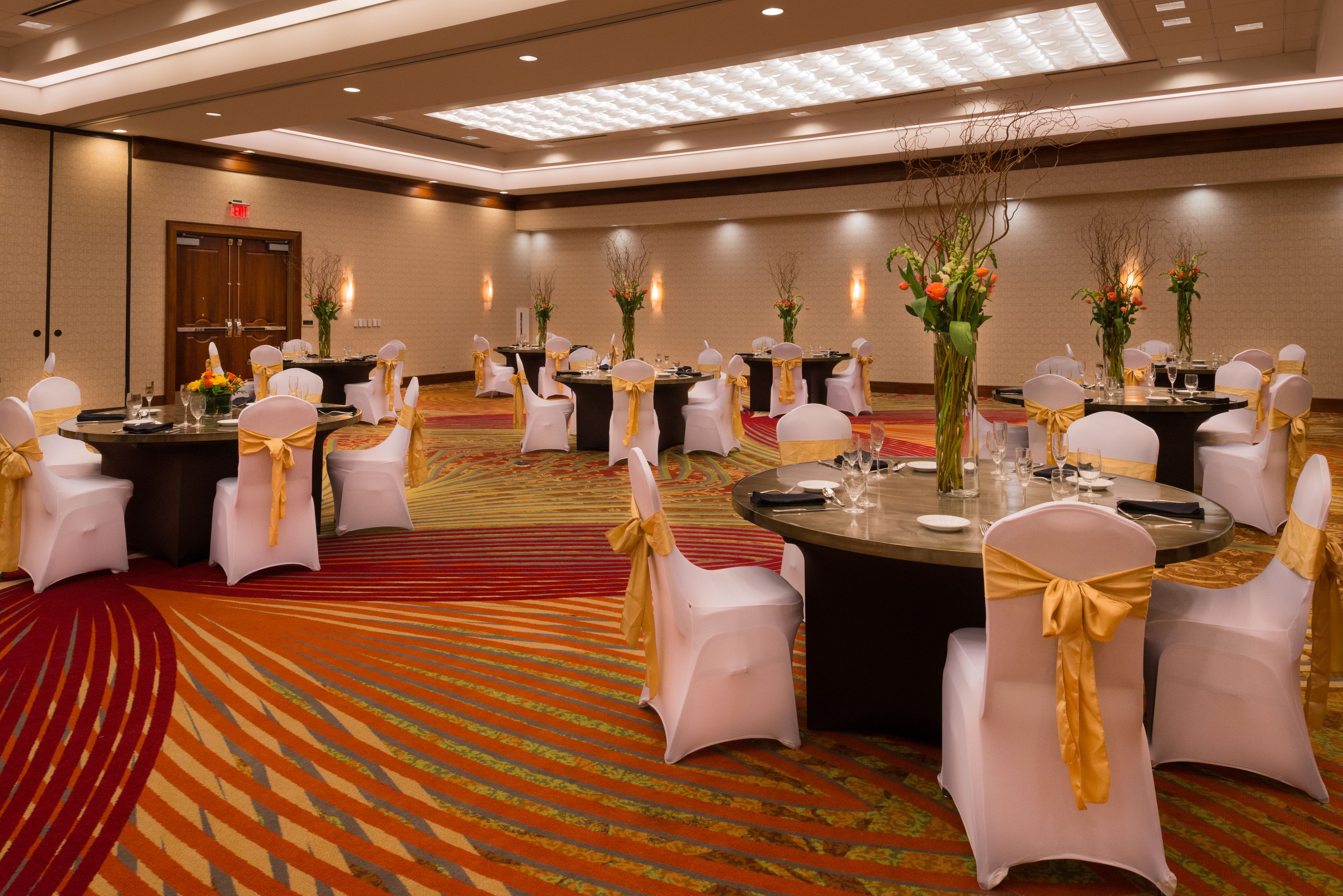 A banquet hall with round tables, white chairs with gold sashes, and floral centerpieces.