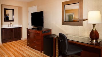 A hotel room with a wooden vanity, a flat-screen TV, a desk with a chair, and a lamp.