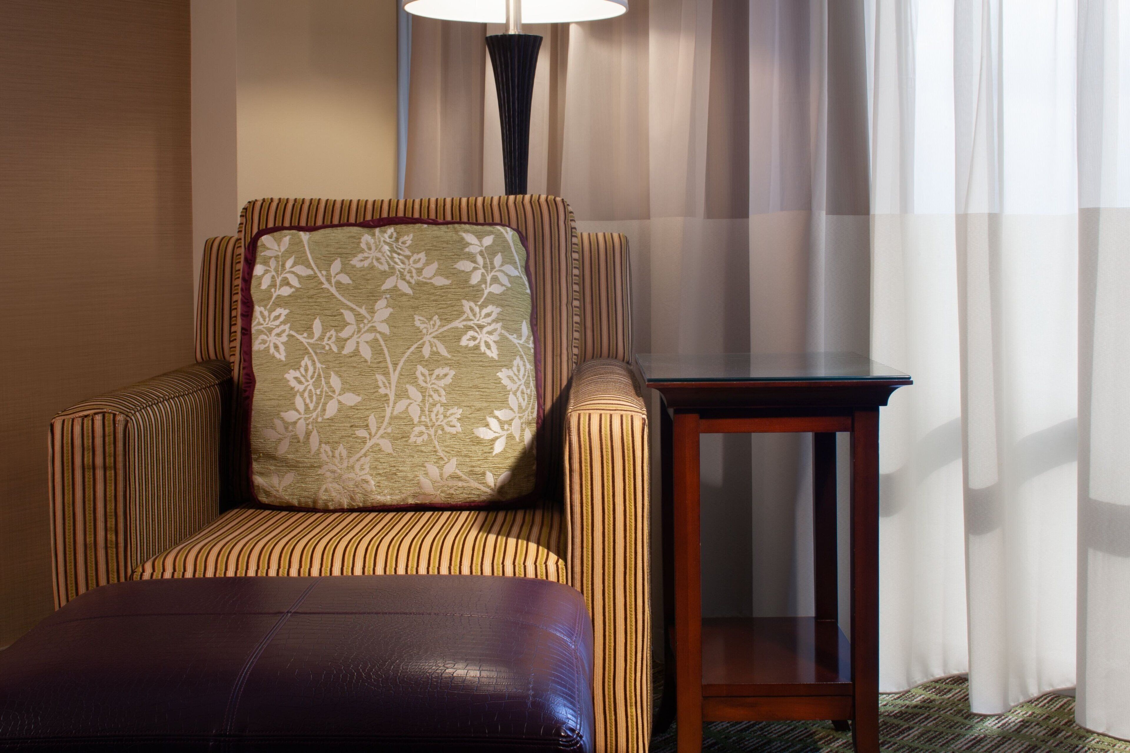 A striped armchair with a floral pillow, a small wooden side table, and a leather ottoman in a hotel room.