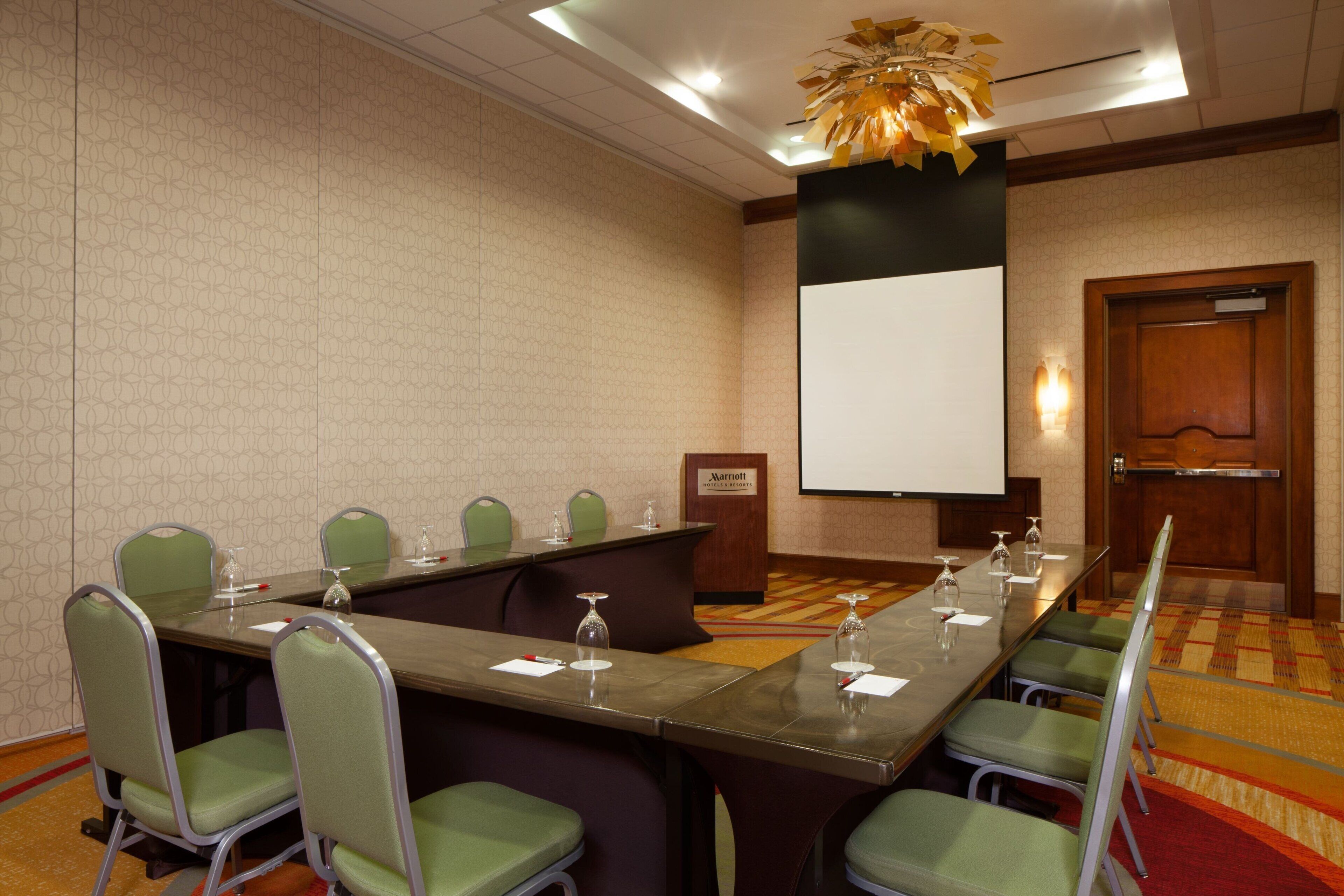 A conference room with a long table, green chairs, a podium, and a projector screen.