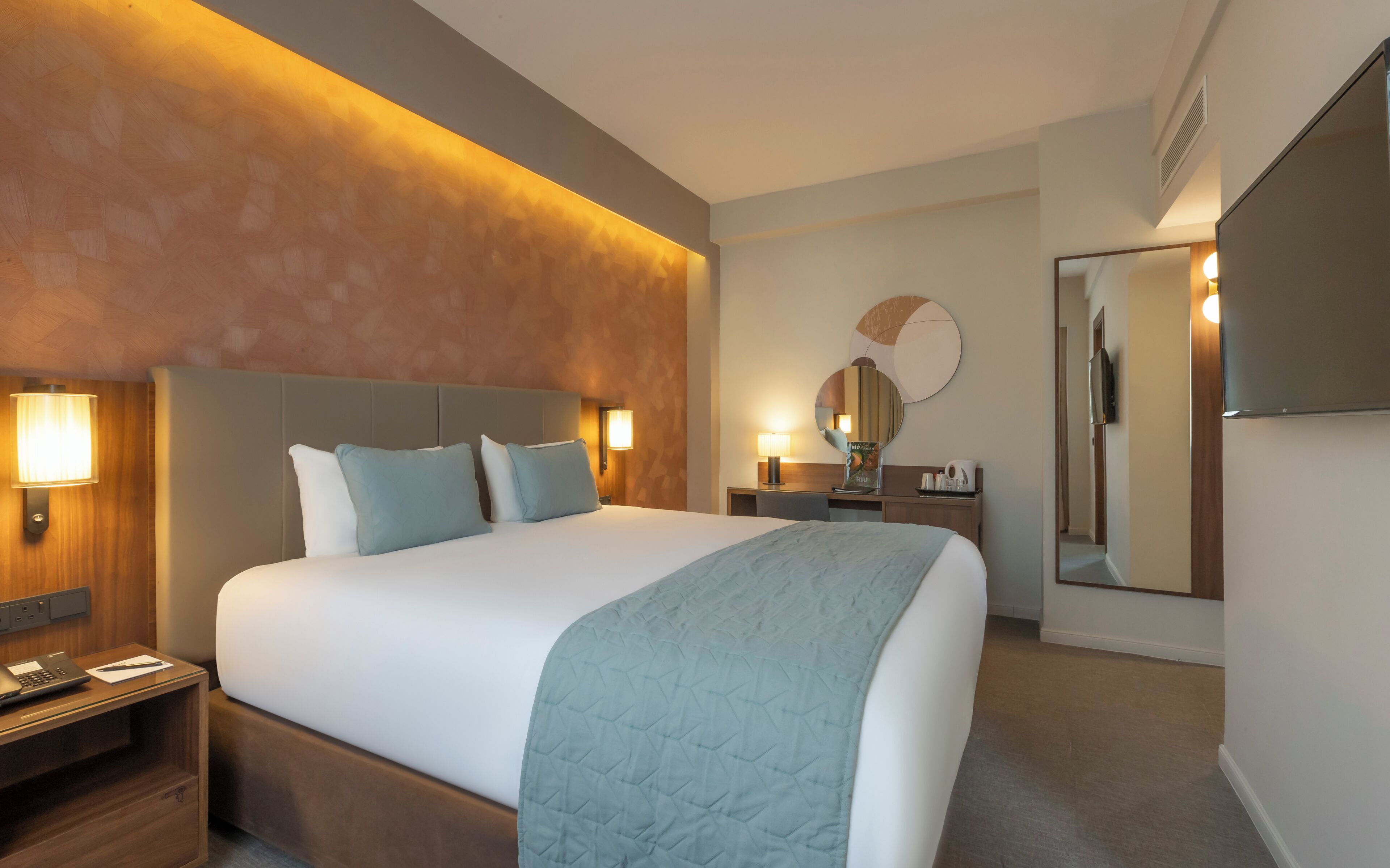 A hotel room with a large bed, bedside lamps, a desk with a mirror, and a flat-screen TV.