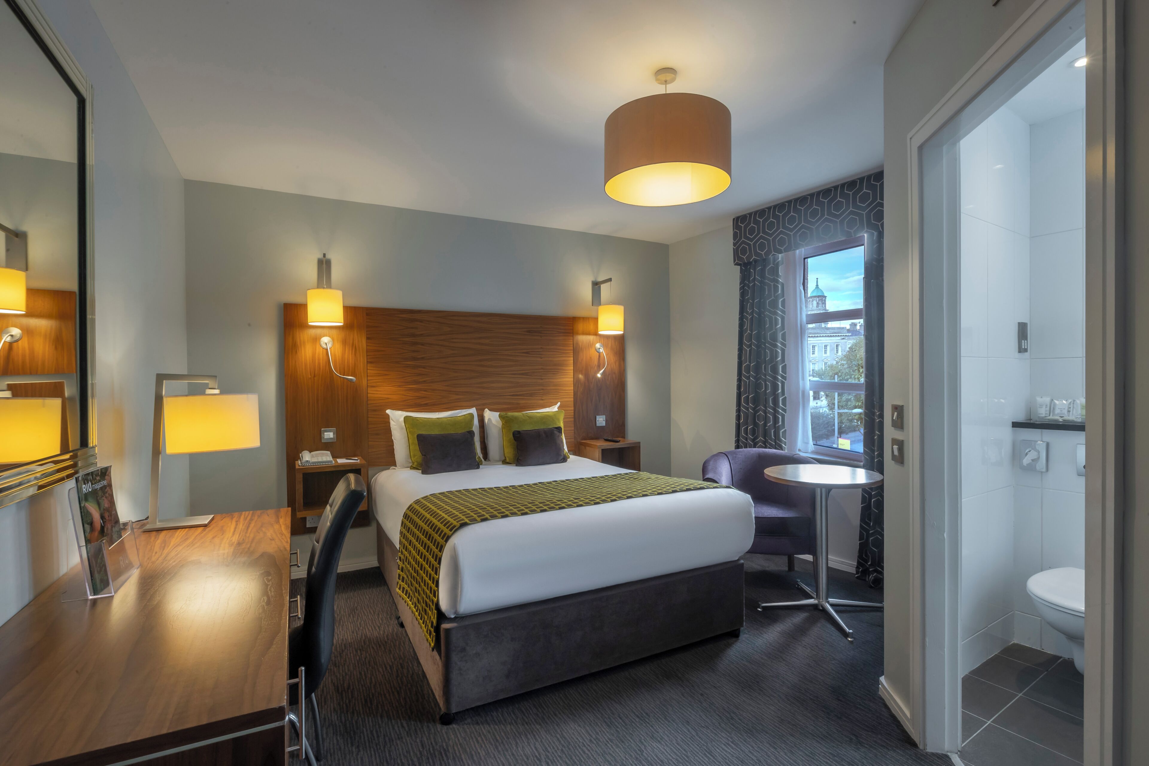 Une chambre d’hôtel avec un lit, des lampes de chevet, un bureau, une chaise, une table ronde et une vue sur un immeuble par la fenêtre.