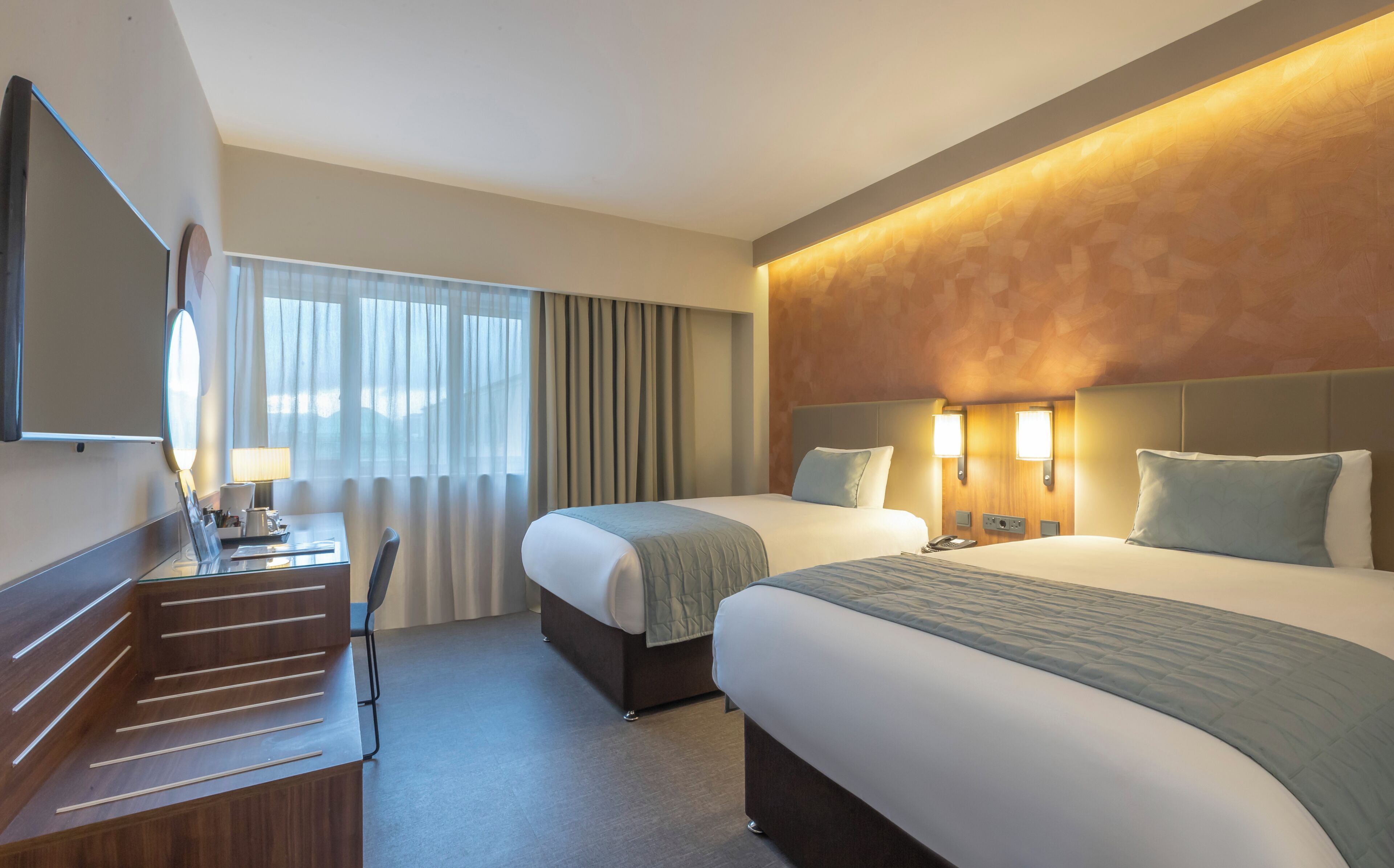 Une chambre d’hôtel avec deux lits, un bureau avec une chaise, un miroir et une fenêtre avec des rideaux.