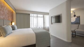 Habitación de hotel con cama, televisor, sofá y un amplio ventanal con cortinas.