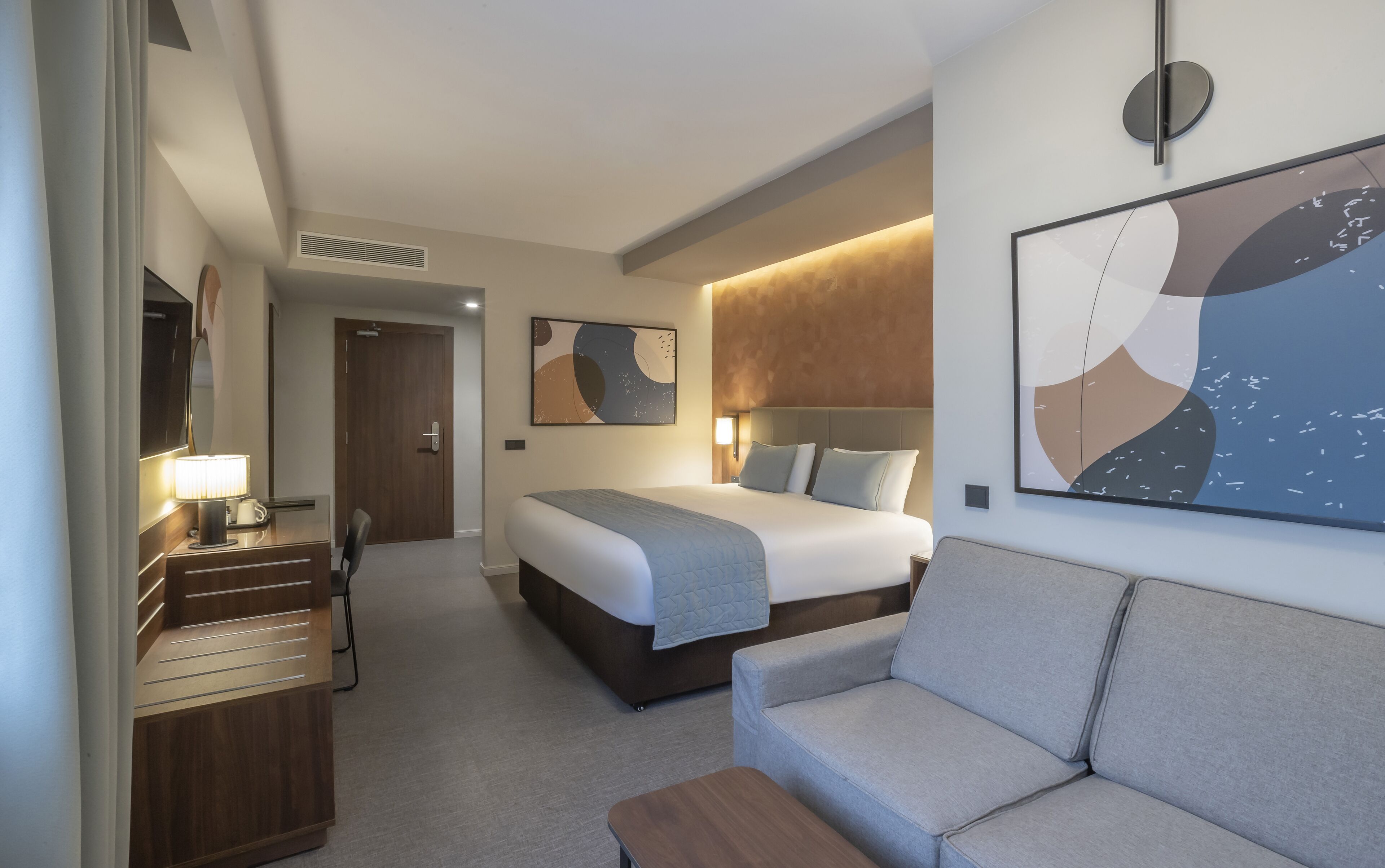 A hotel room with a bed, a sofa, a desk with a lamp, and artwork on the walls.