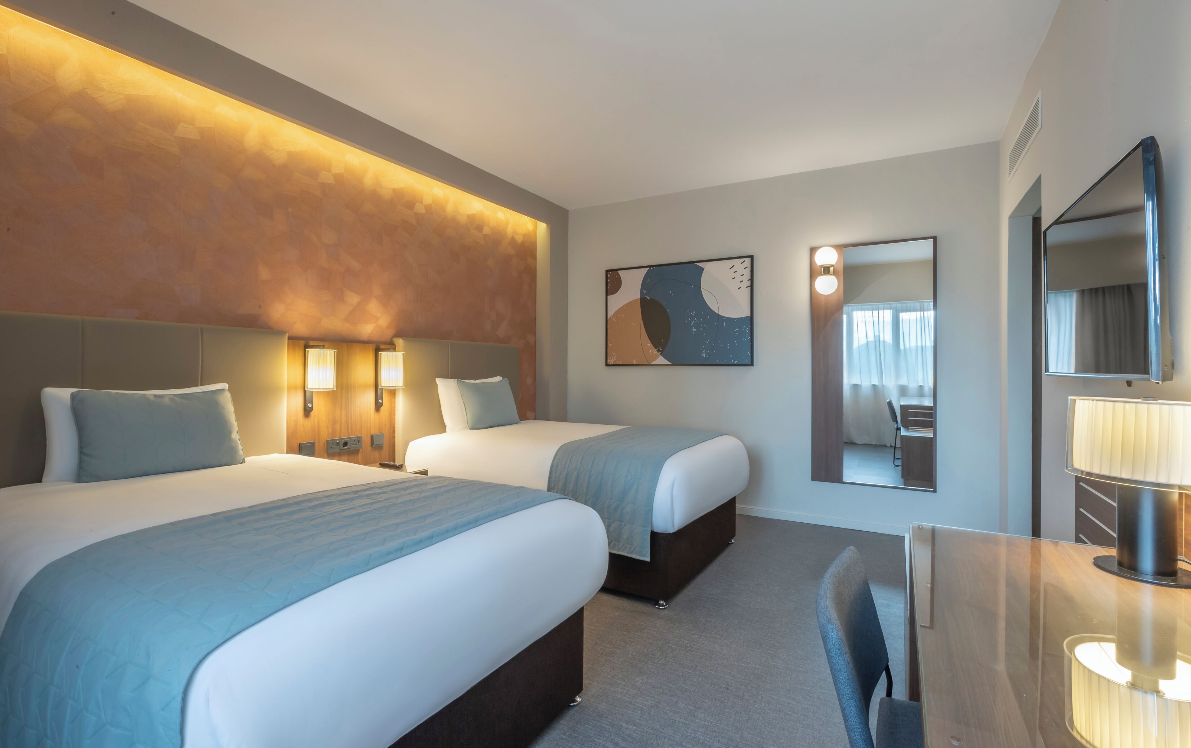 A hotel room with two beds, a desk, a chair, a mirror, and a painting on the wall.