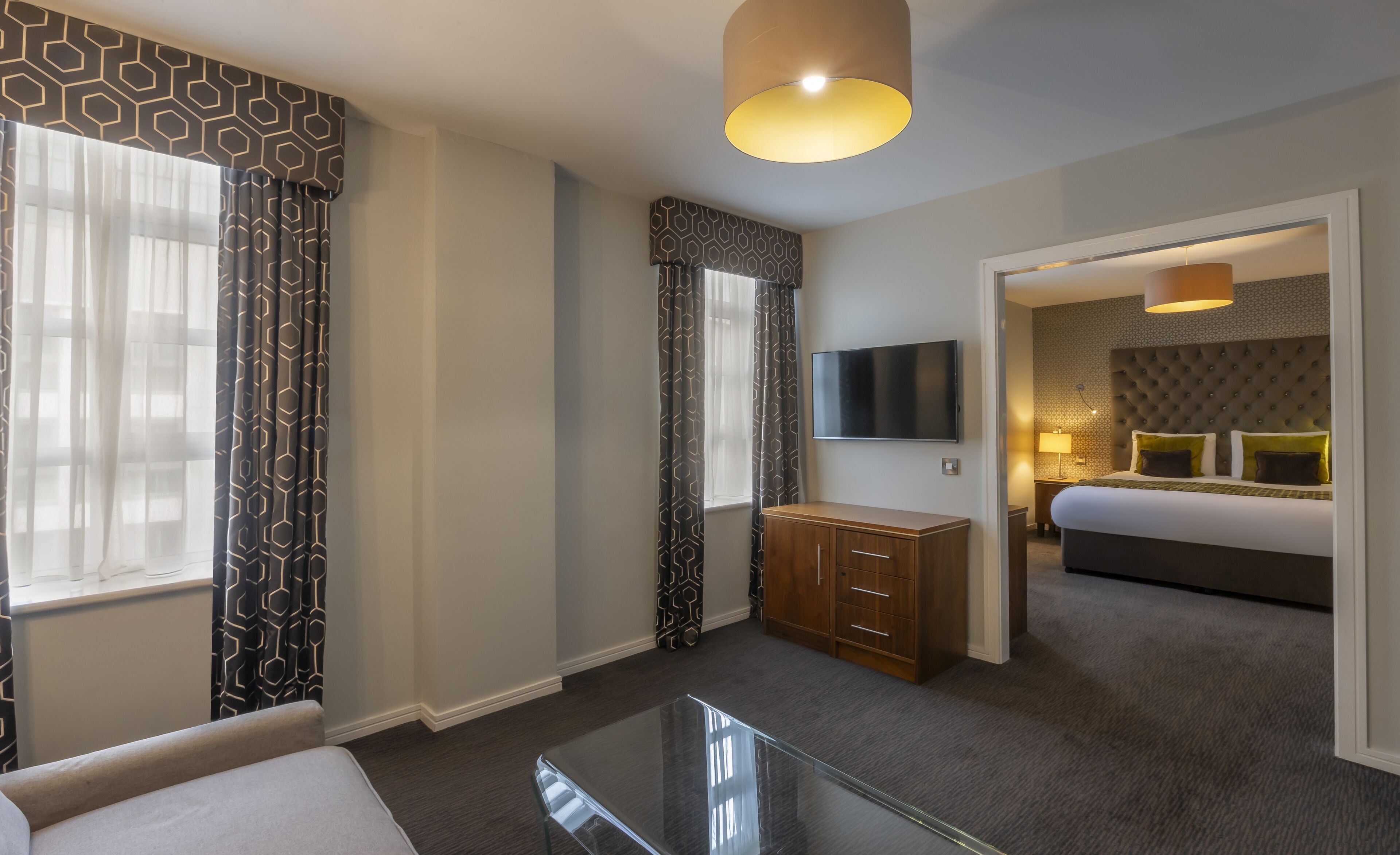 Una habitación de hotel con una mesa de centro de vidrio, un televisor montado en la pared, una cama con cabecera capitonada y un armario.