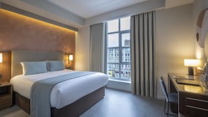 A modern hotel room with a large bed, a desk, and a view of the city through the window.