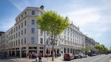 A large, multi-story hotel with a classic architectural style, featuring a prominent corner entrance and a row of shops on the ground floor.