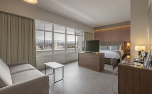 A hotel room with a bed, a TV, a desk, a chair, and large windows offering a city view.