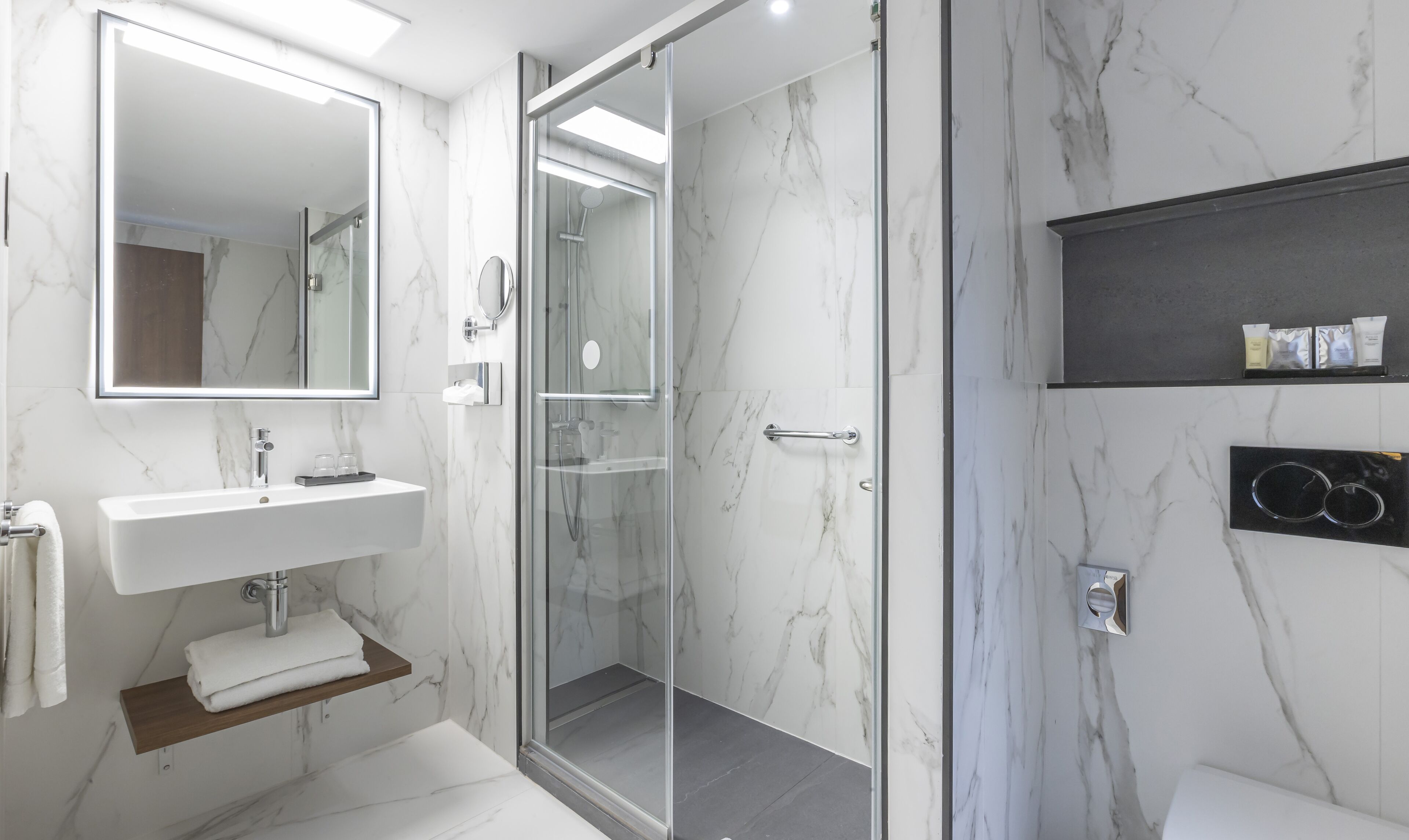 A modern bathroom with a glass-enclosed shower, a white sink with a marble countertop, and a mirror above it. There are towels on a shelf and a small black shelf with toiletries.