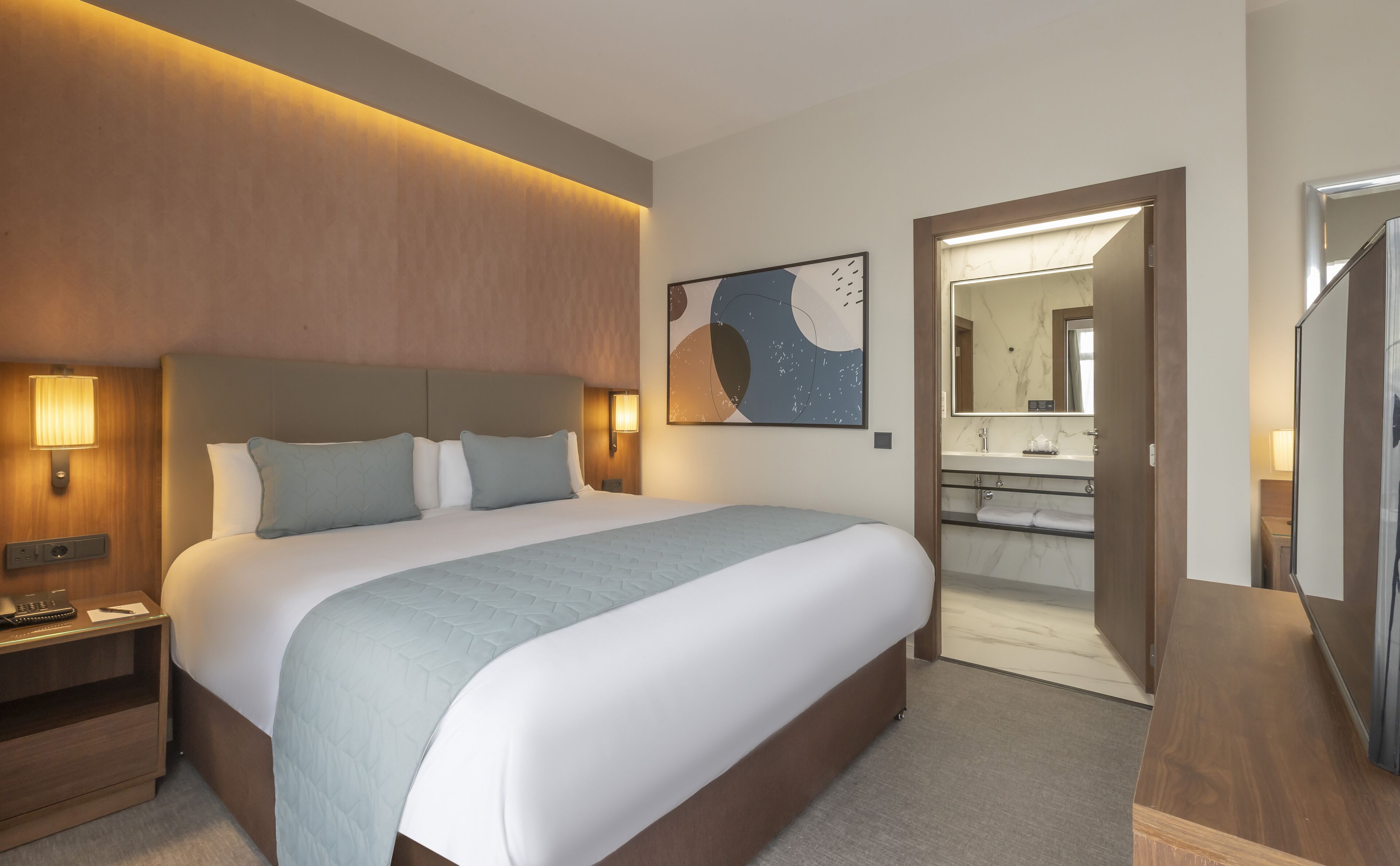 A hotel room with a large bed, bedside lamps, a nightstand, a bathroom with a mirror, and a wooden floor.