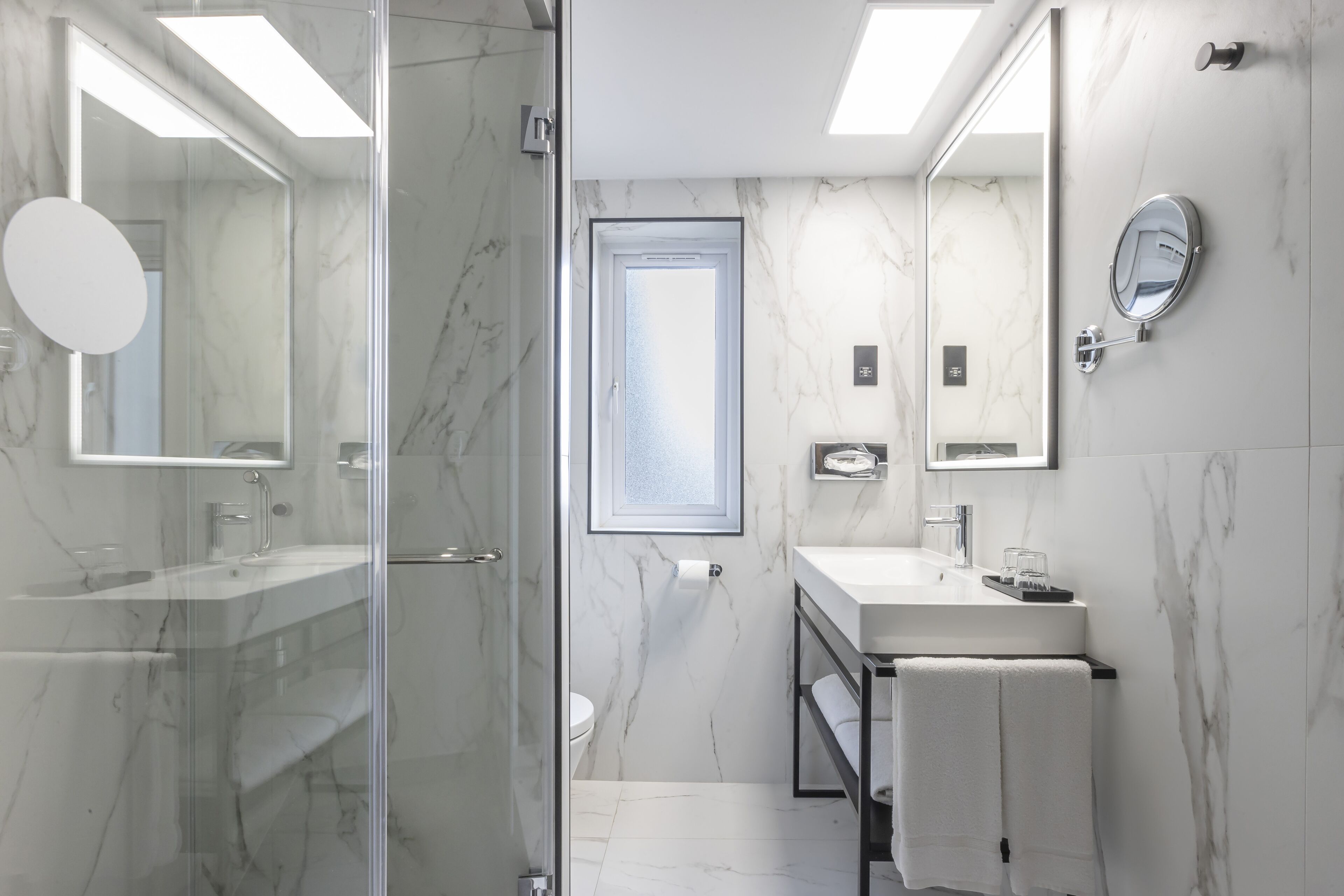 Un baño moderno con cabina de ducha de vidrio, un lavabo blanco con espejo y pared revestida de mármol.