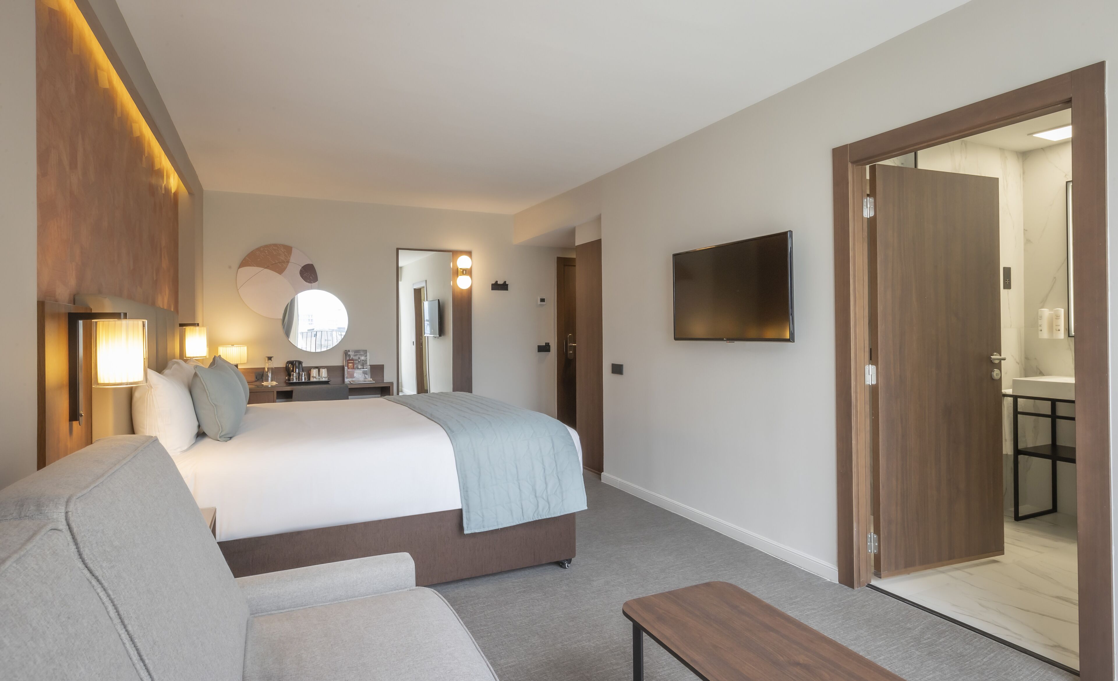 Quarto de hotel com cama, sofá, televisão, escrivaninha com abajur e banheiro visível através de uma porta aberta.