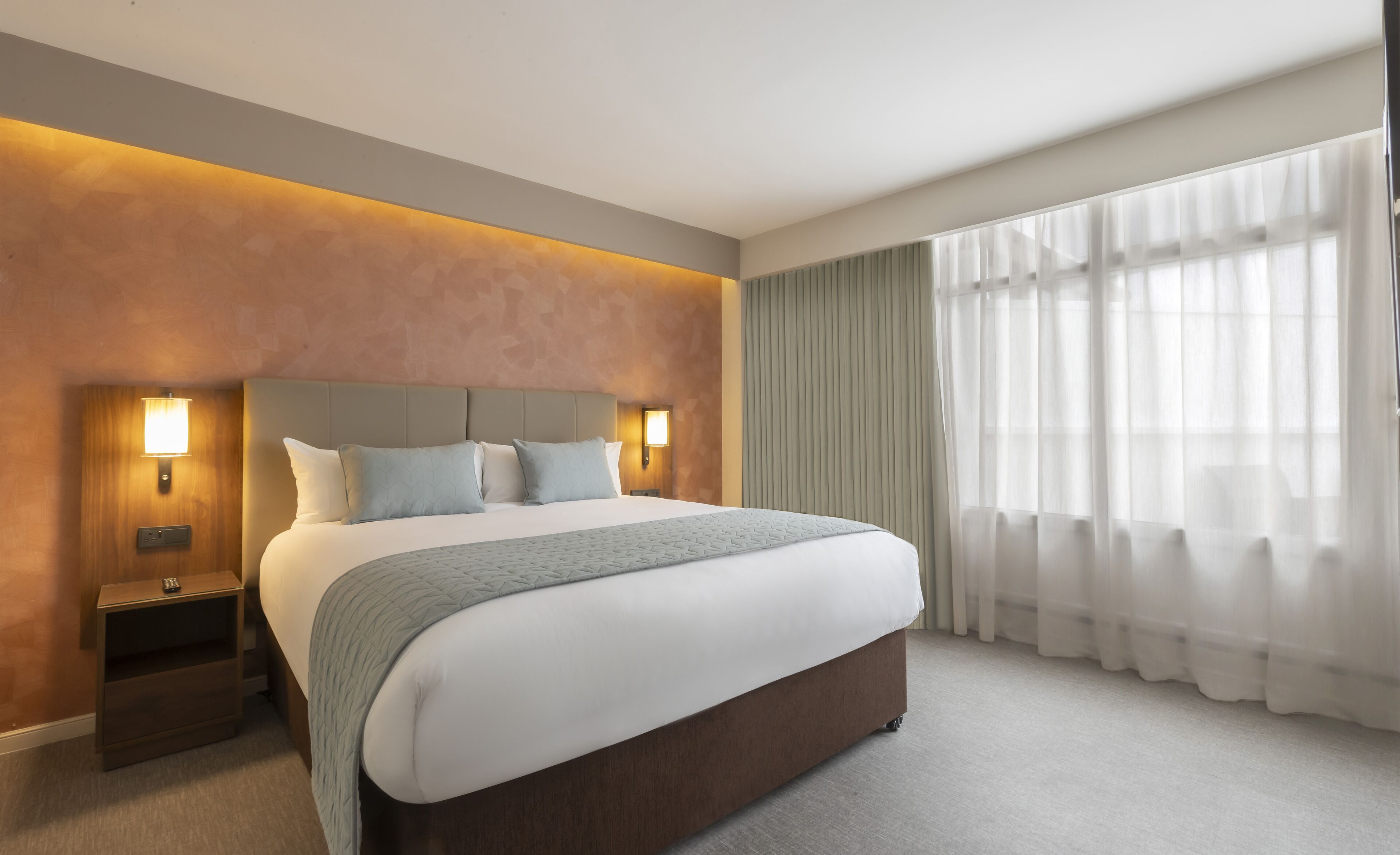 A modern hotel room with a large bed, two bedside tables with lamps, a view of the city through sheer curtains, and a textured wall.