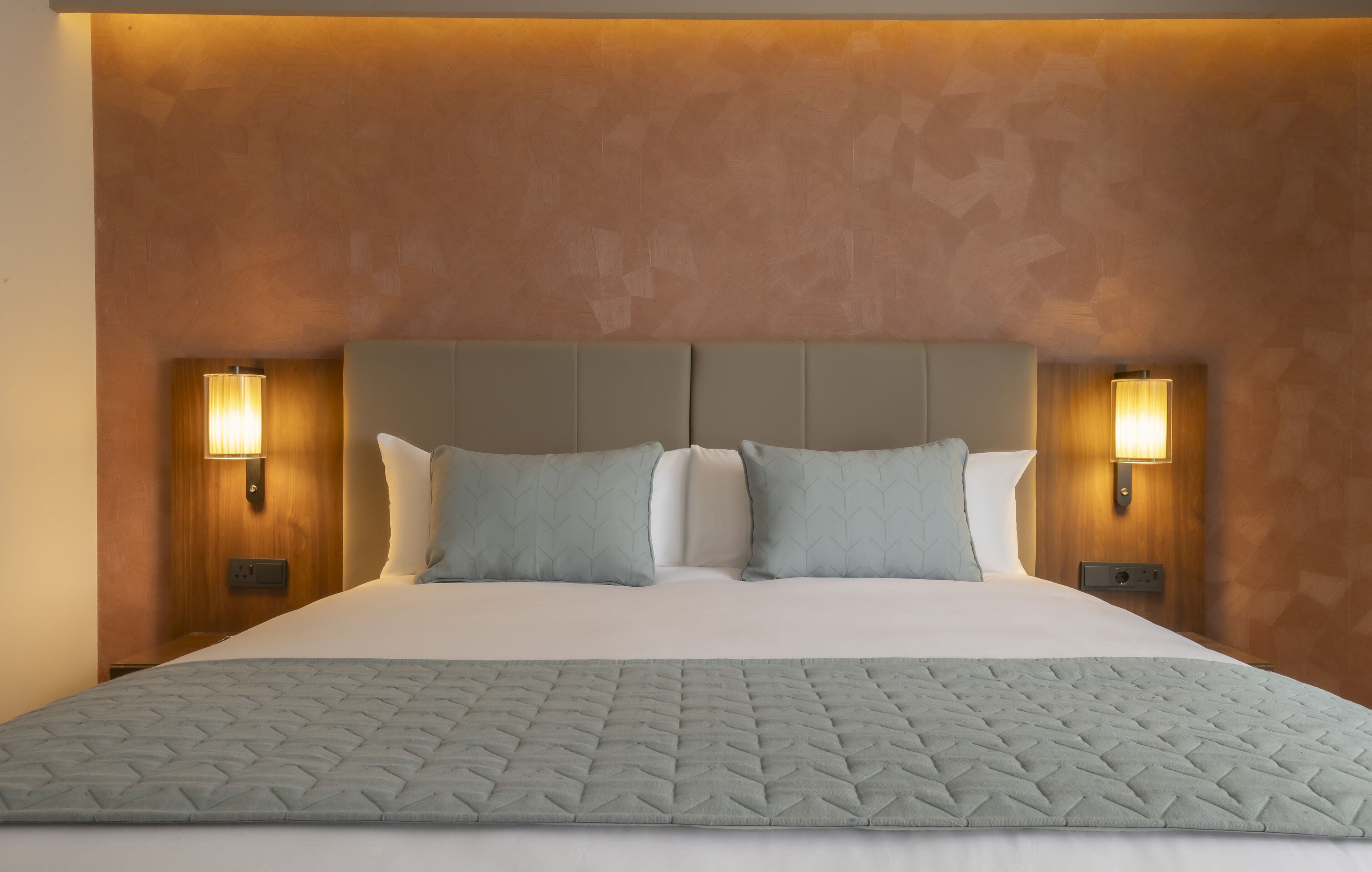 Una cama grande con cubrecolcha acolchado, dos lámparas fijadas a la pared y una pared texturizada detrás de la cama.
