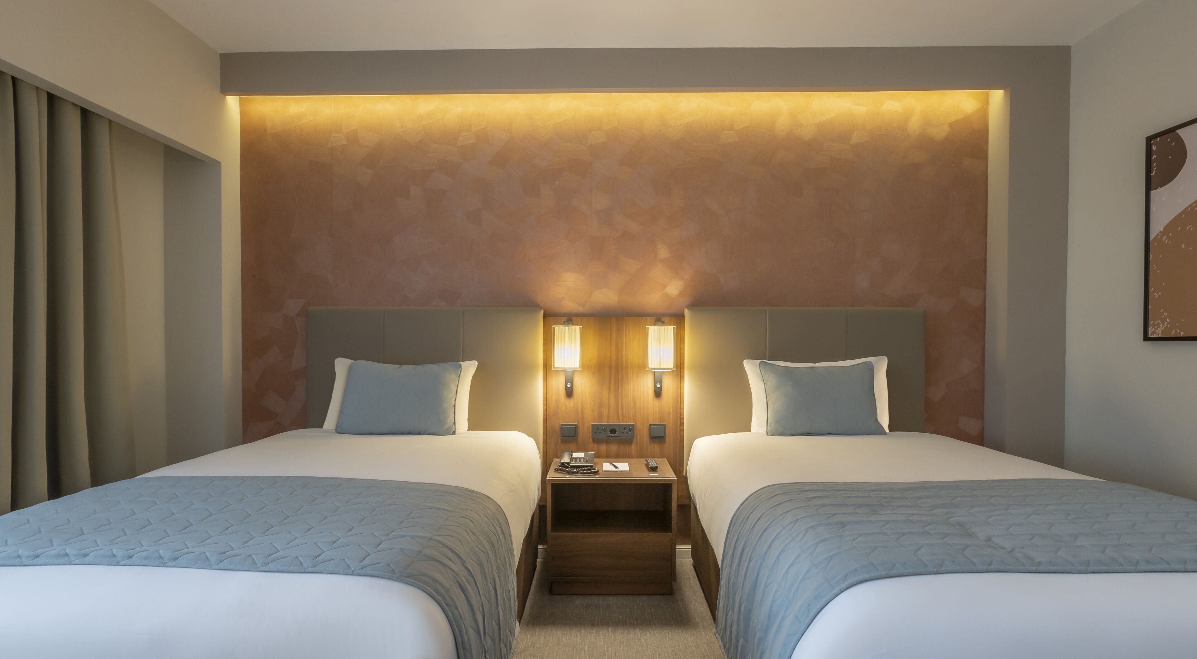 A hotel room with two beds, a nightstand between them, and a wall-mounted lamp.