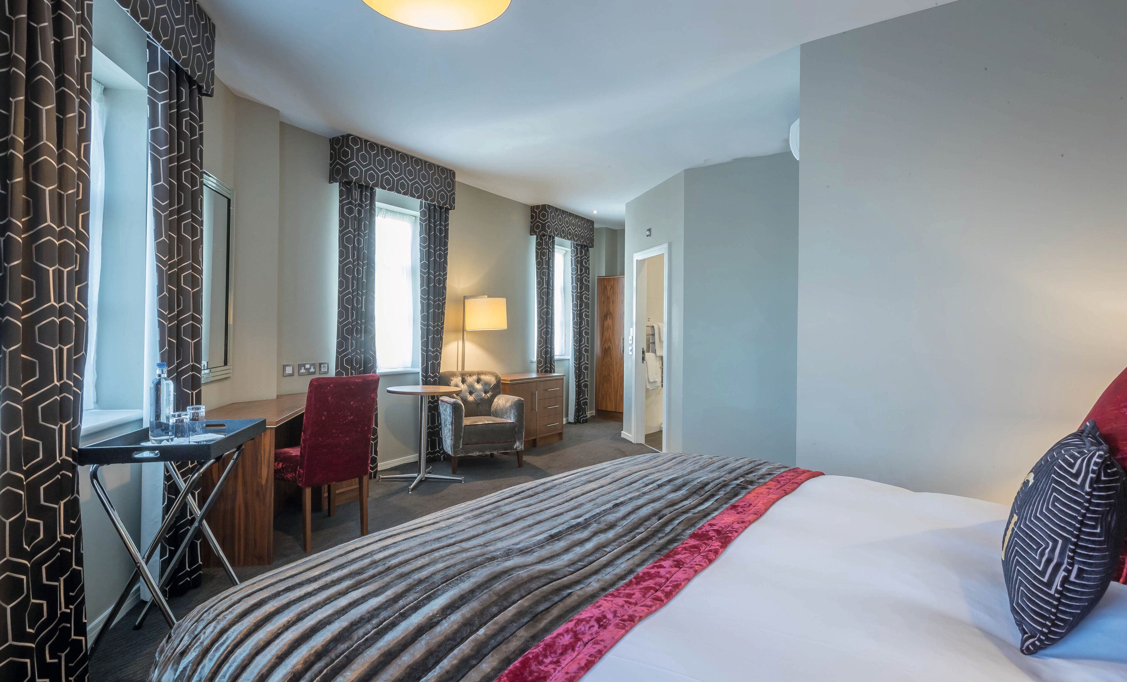 Una habitación de hotel con cama, escritorio, silla, armario y ventana con cortinas.