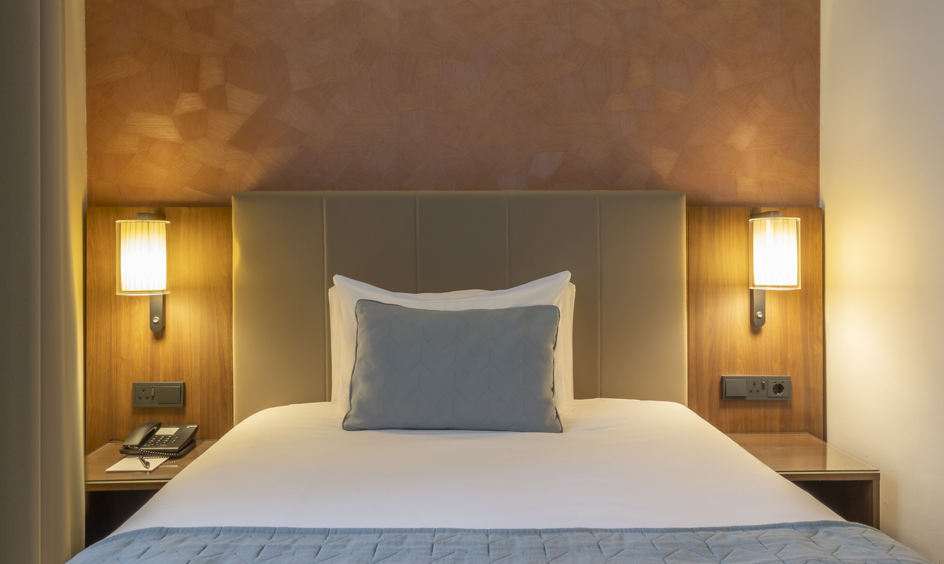 A hotel room with a large bed, two bedside lamps, a nightstand with a phone, and a wall socket.