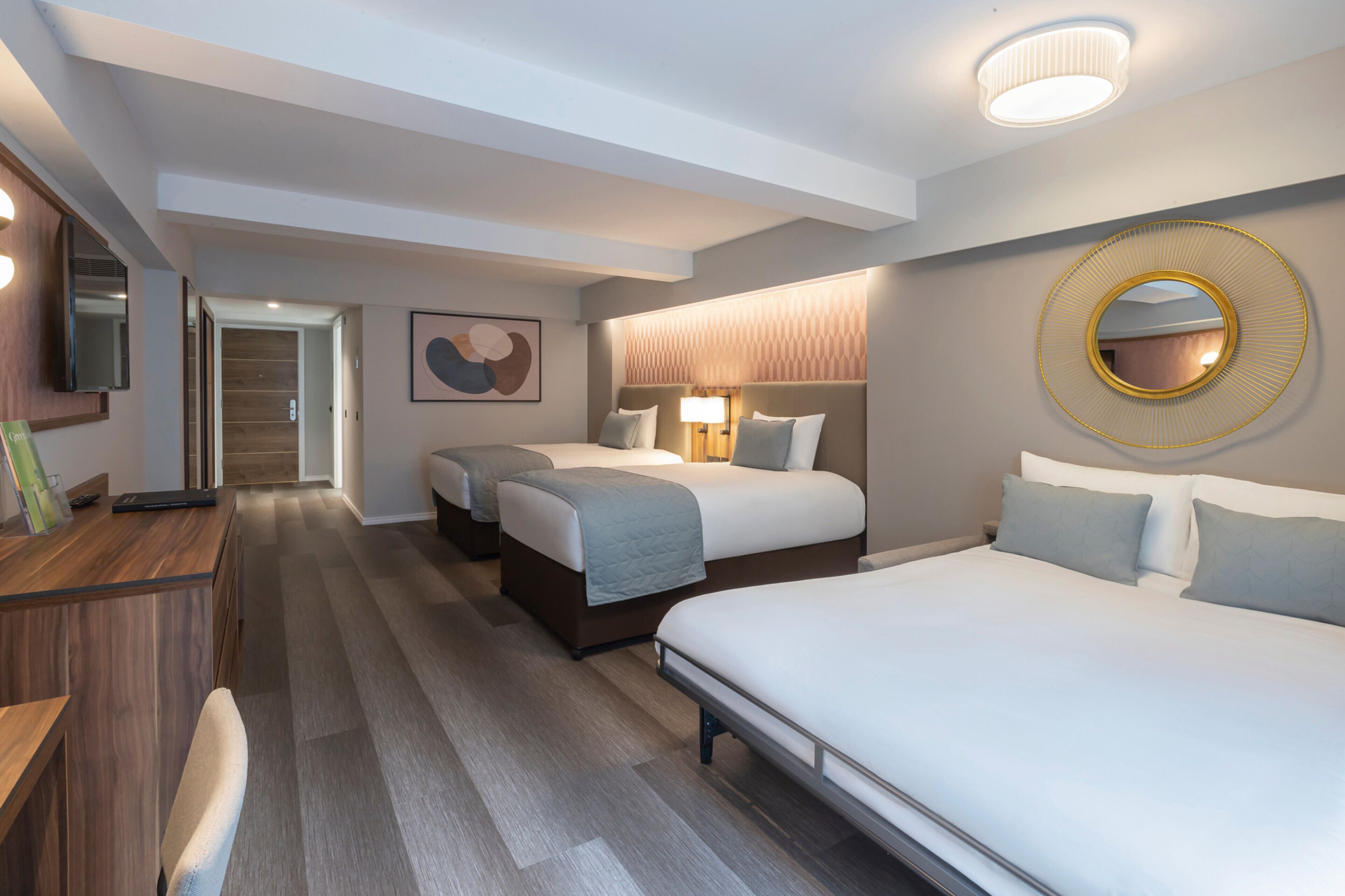 Une chambre d’hôtel avec trois lits, un bureau, un miroir et une lampe fixée au mur.